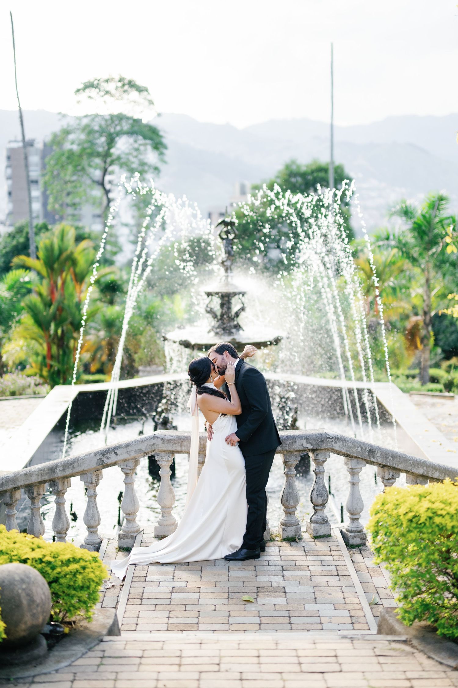 Elegant couple posing by ornate fountains and stone balustrades in a tropical garden.