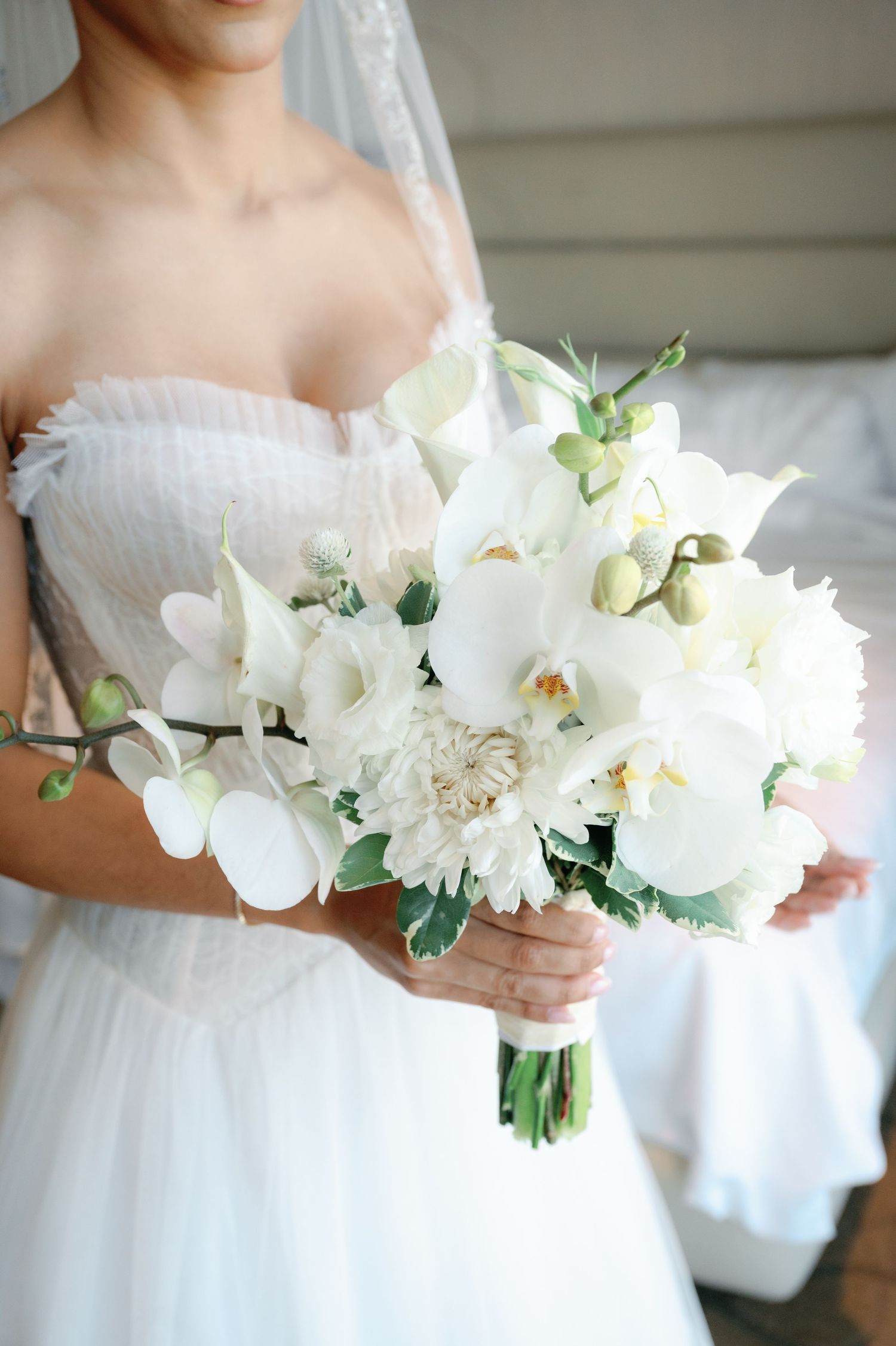 A stunning white bridal bouquet featuring orchids, dahlias and delicate greenery against a white wedding dress.