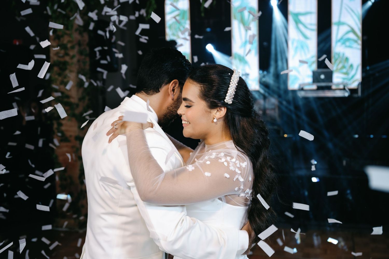 A couple dances together at a wedding reception as confetti falls around them in a dimly lit room.