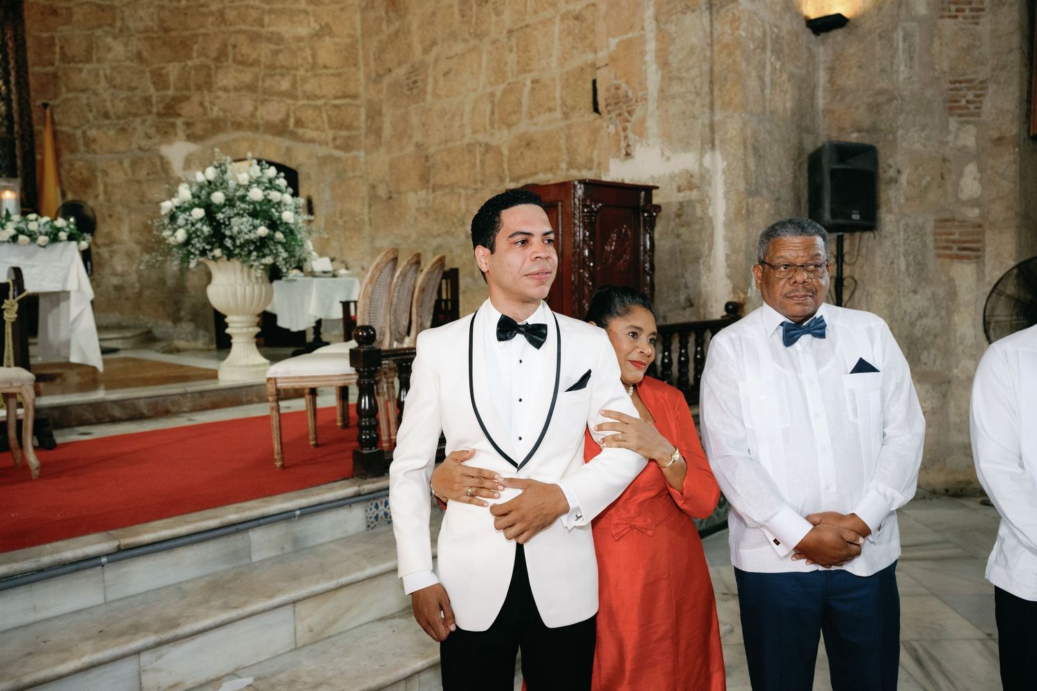 A group in formal attire including white tuxedo jackets and bow ties gather at a stone-walled venue during a celebration.