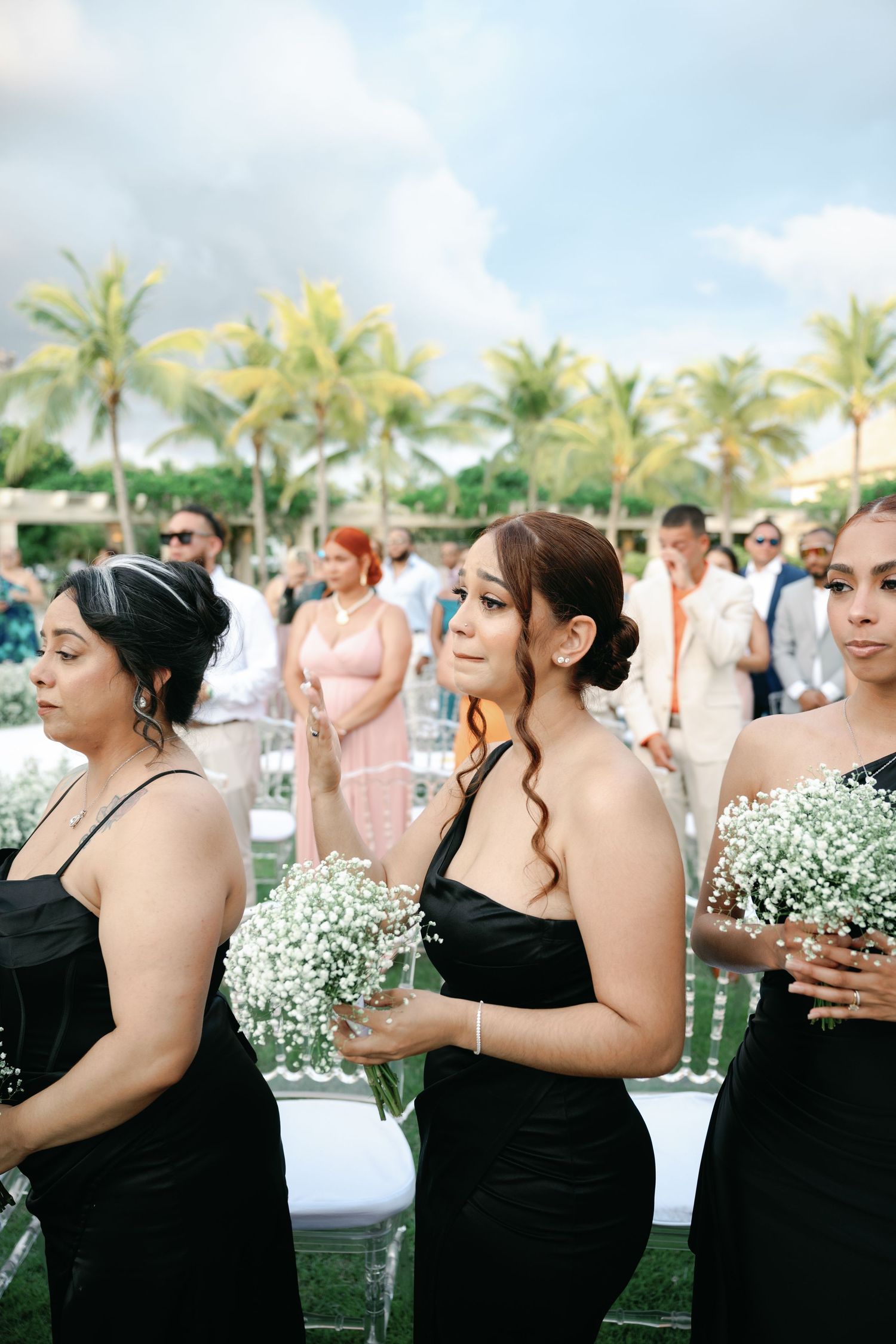 Bridesmaids in black dresses holding white floral bouquets stand in line at an outdoor tropical wedding venue.