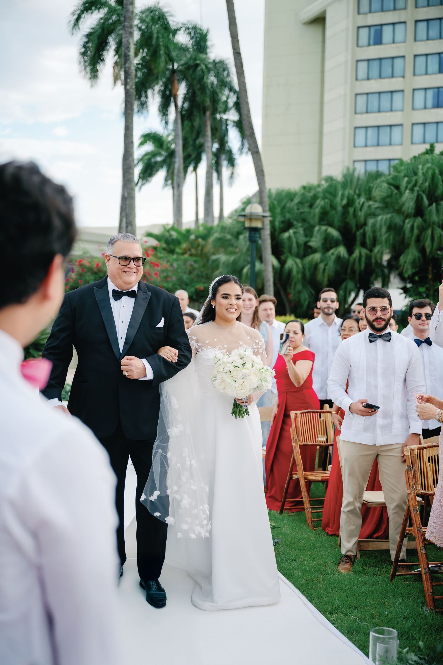 Wedding guests seated in wooden chairs on a lawn with palm trees and a building in the background.
