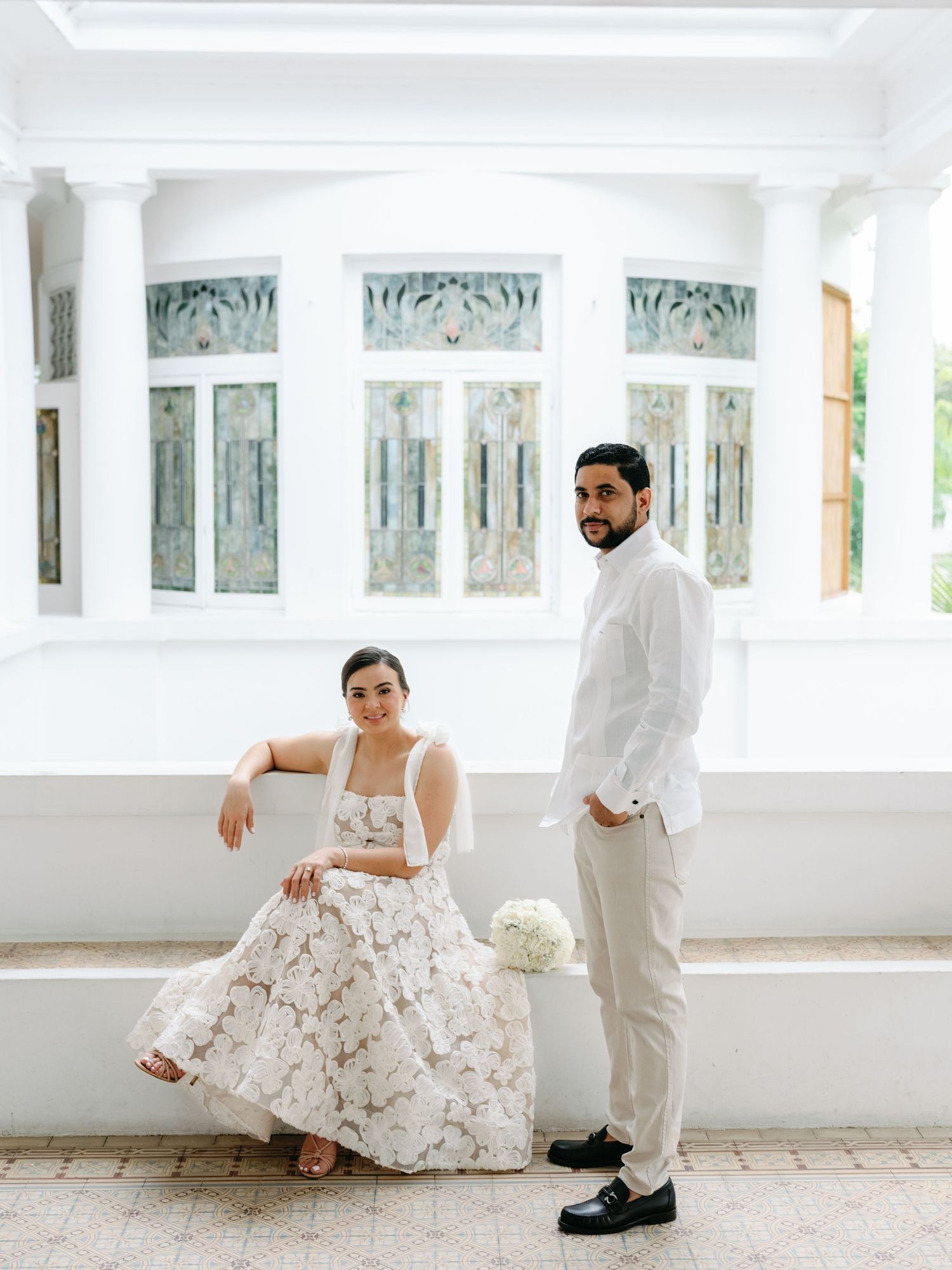A couple in white attire poses elegantly in a bright sunlit veranda with ornate windows and columns.
