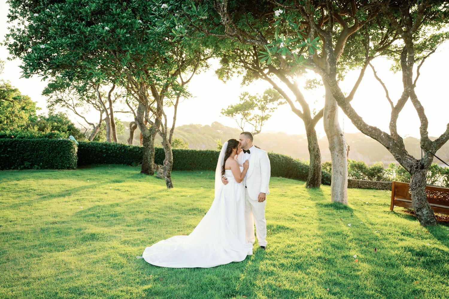 Bride in a flowing white gown embraces her groom on a lush green lawn beneath sunlit trees during a golden hour outdoor wedding.