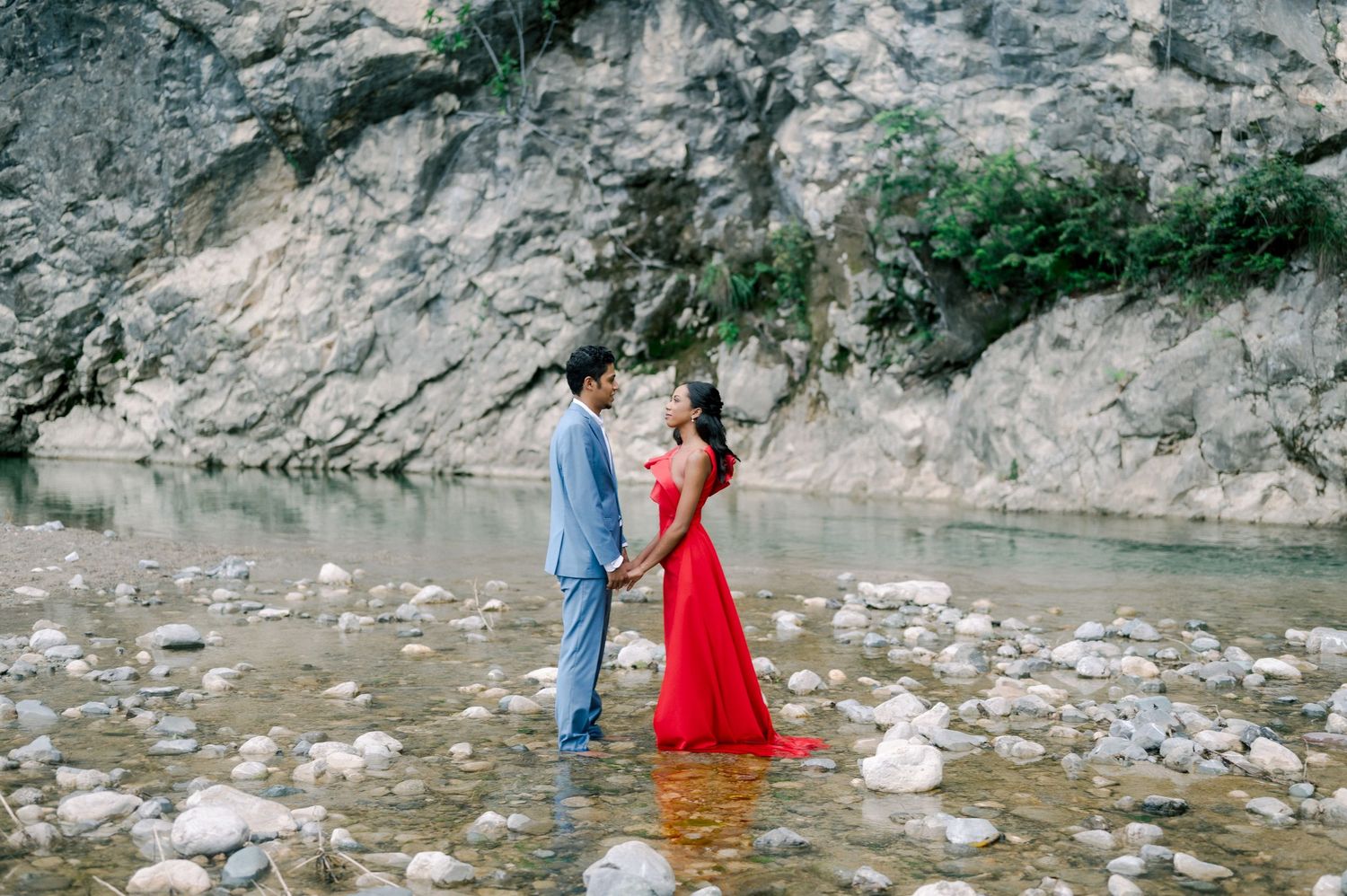 A couple stands together on rocky riverbank with limestone cliffs in background as woman wears flowing red dress.