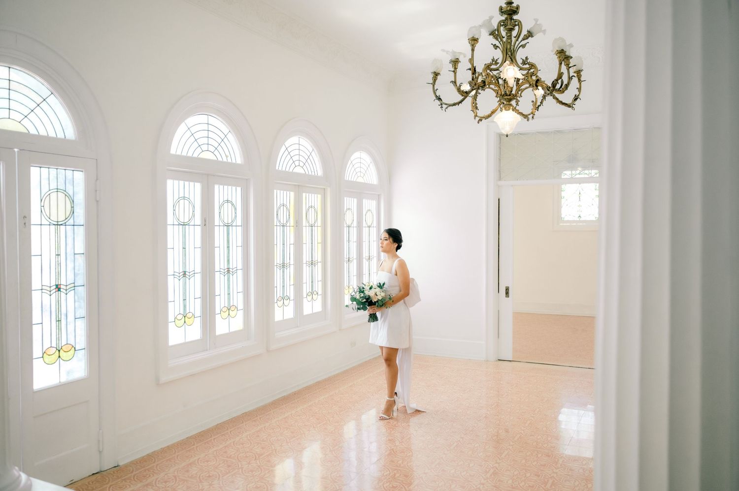 A bright white ballroom with arched windows, chandelier, and natural light streaming through the elegant space.
