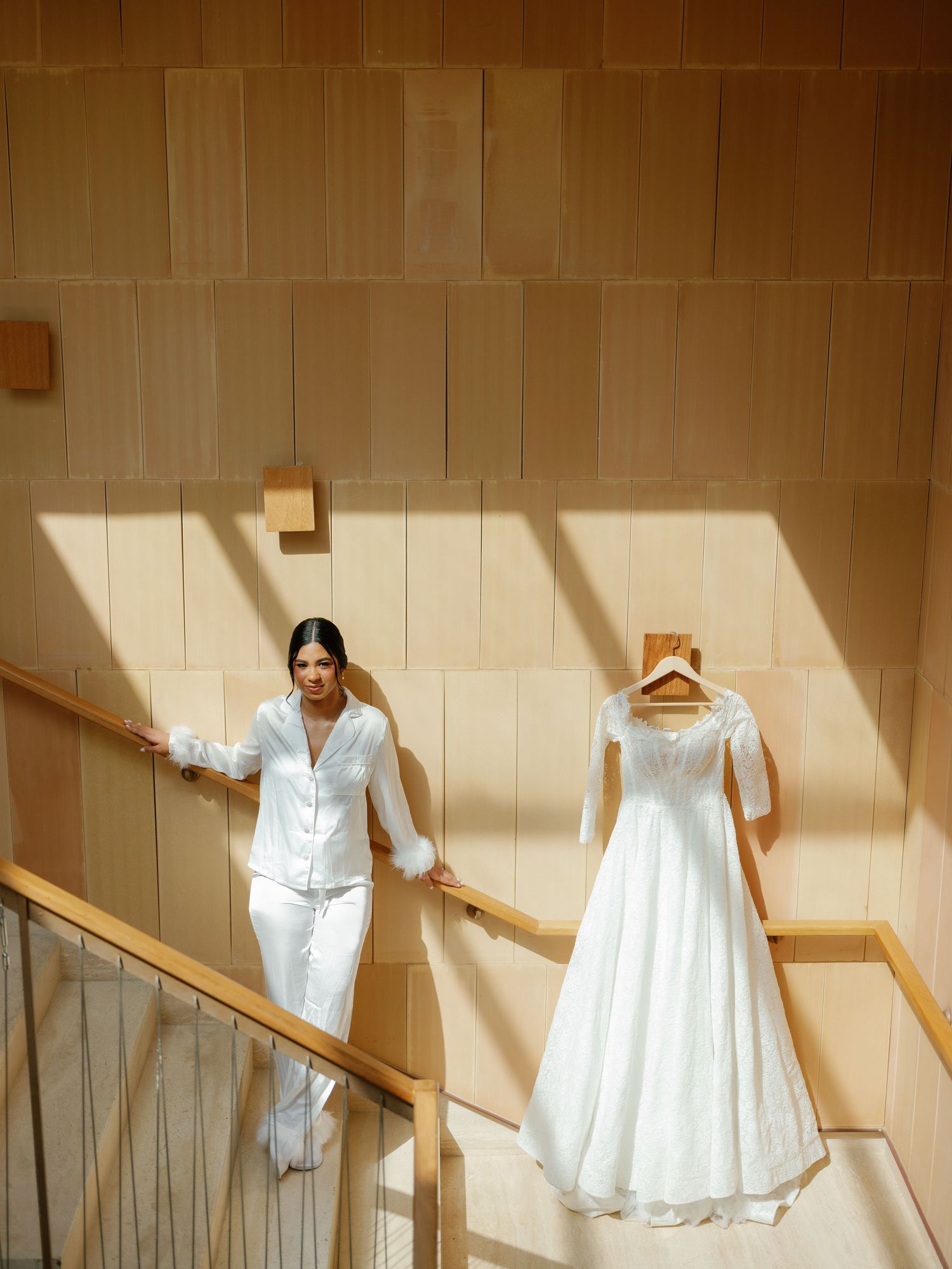 A white wedding dress hangs on display against a beige wall in a sunlit stairwell.