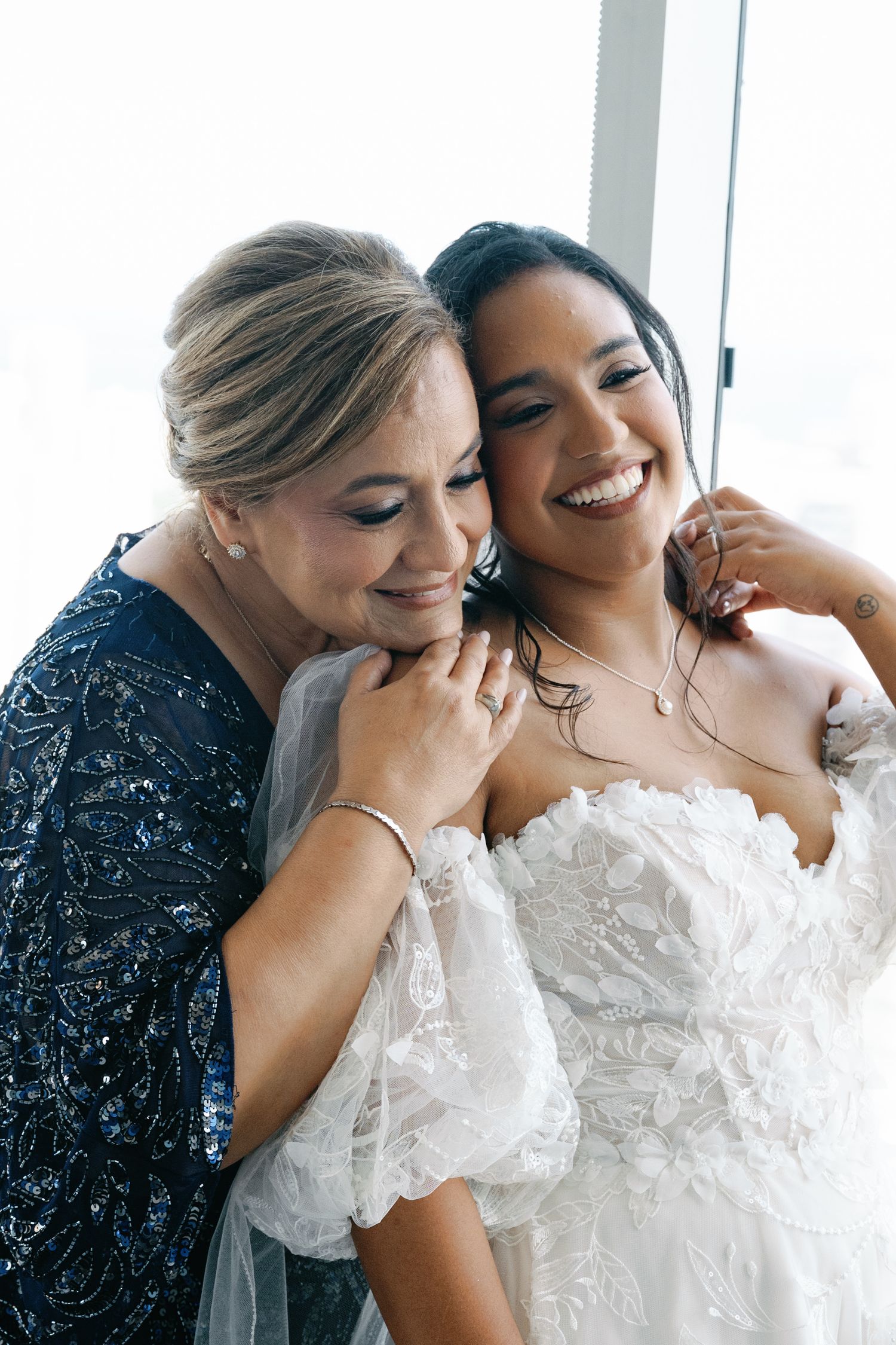 Two women share a joyful moment in formal attire, one wearing a navy beaded dress and the other in a white lace gown.