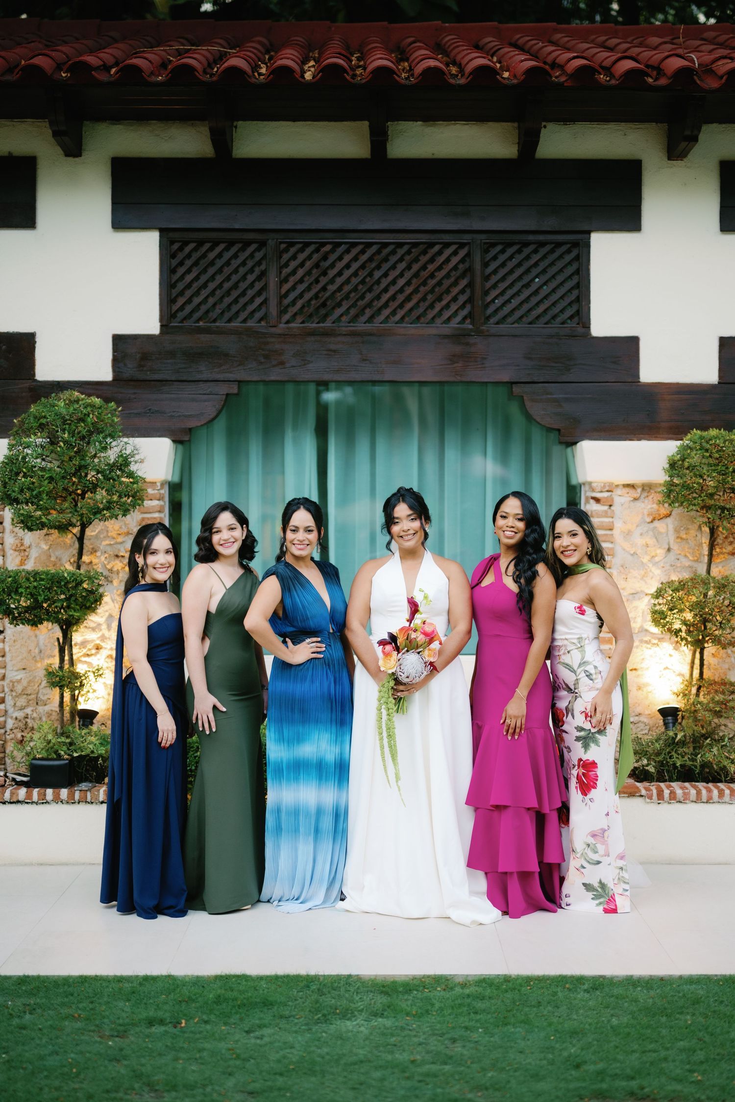Wedding party group poses together in front of white building with teal doors and Spanish tile roof details.