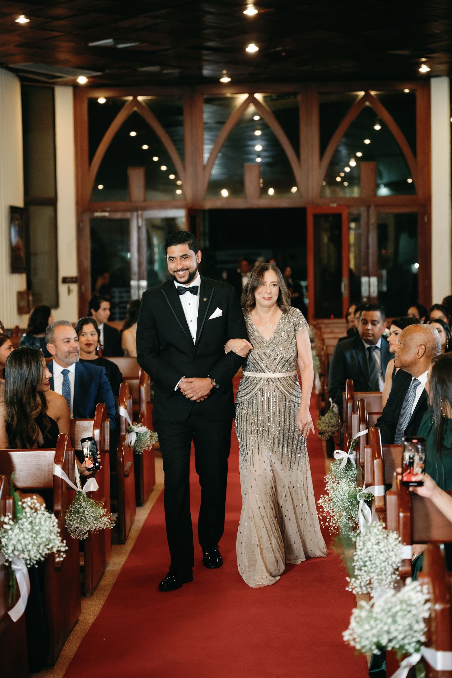 Guests walk down a red-carpeted aisle lined with baby's breath in an elegant venue with arched wooden beams and warm lighting.