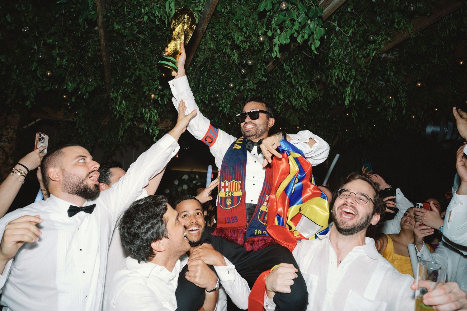 People in white formal attire celebrate enthusiastically at an outdoor party with drinks and dancing.