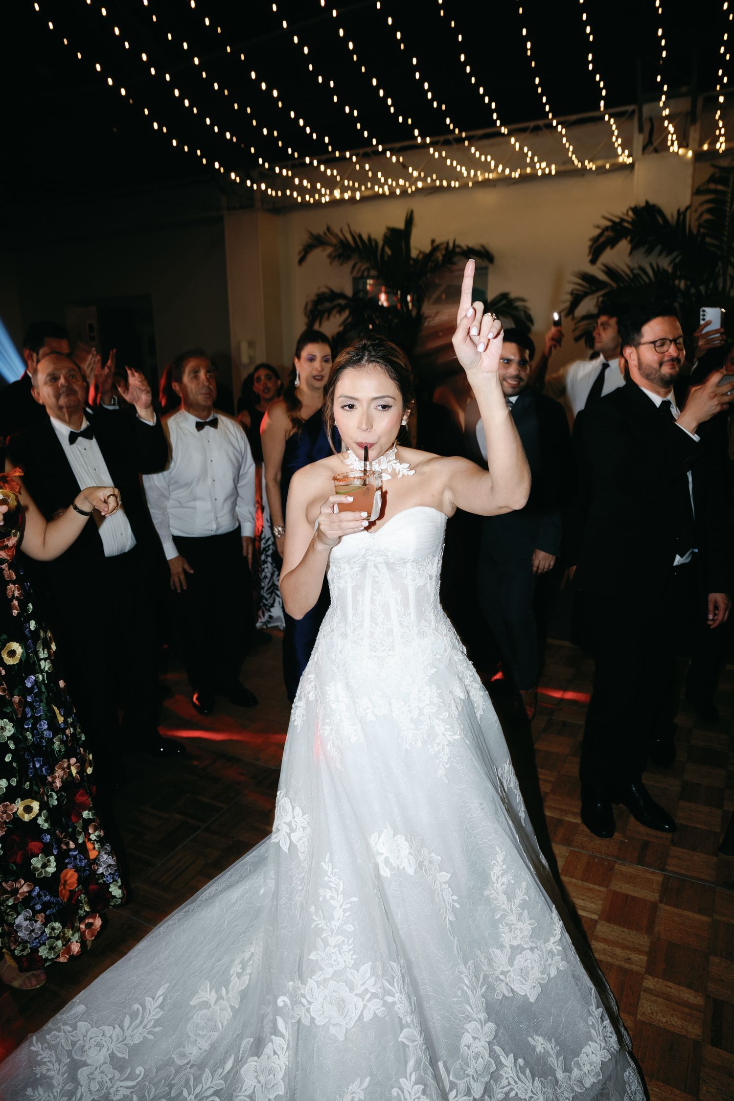 Bride in white lace gown dances under string lights at wedding reception.