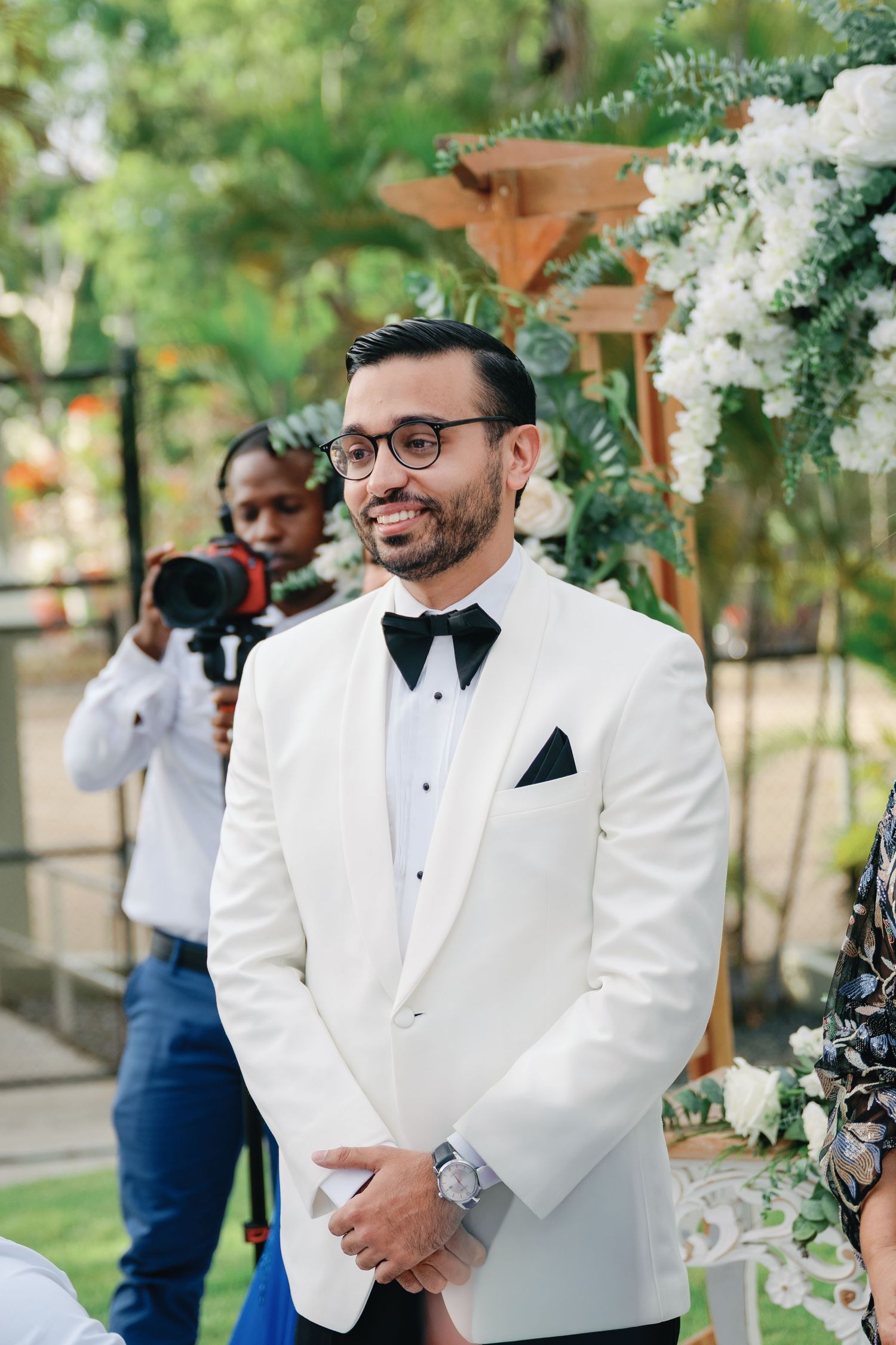 A groom in a white tuxedo jacket and glasses reacts emotionally during an outdoor wedding ceremony.