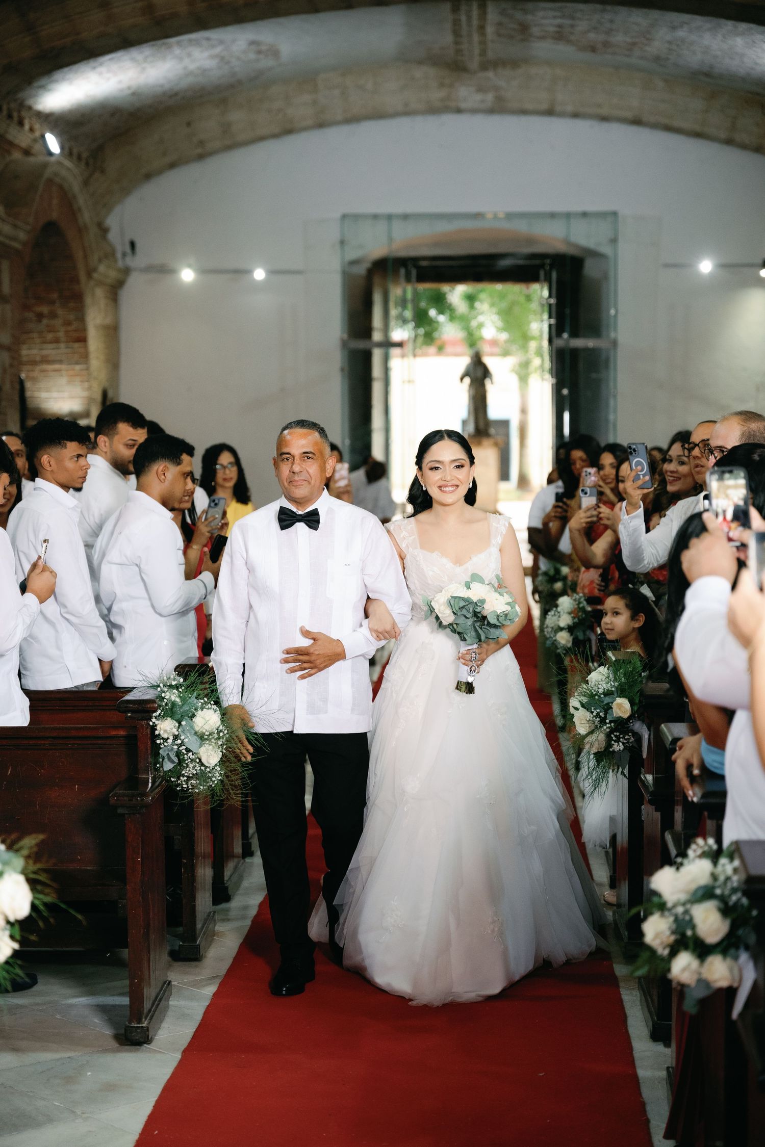A wedding ceremony in a beautiful stone church with a red carpet aisle and guests seated in wooden pews.
