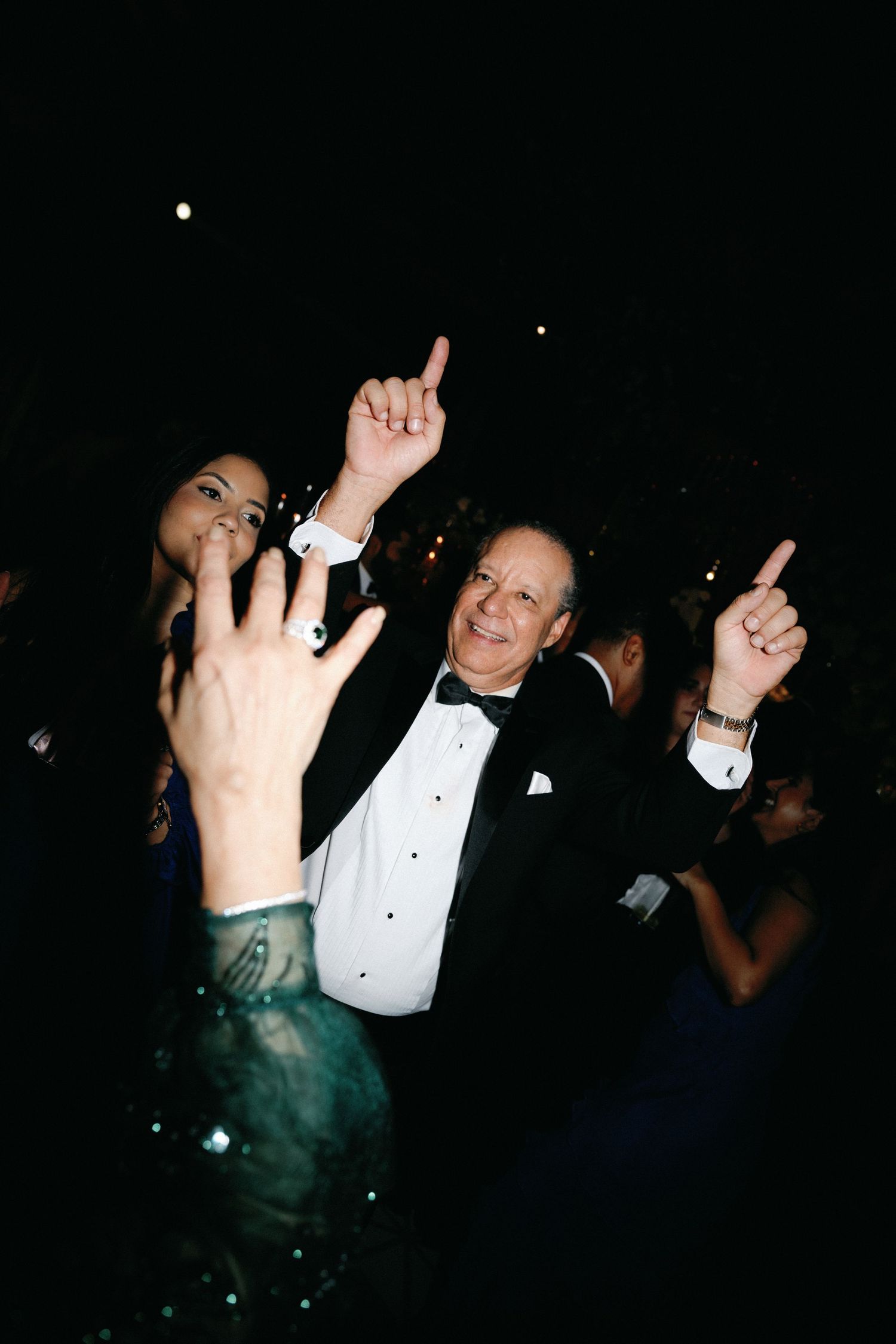 People dancing and raising their hands in formal attire at a dark celebration event.