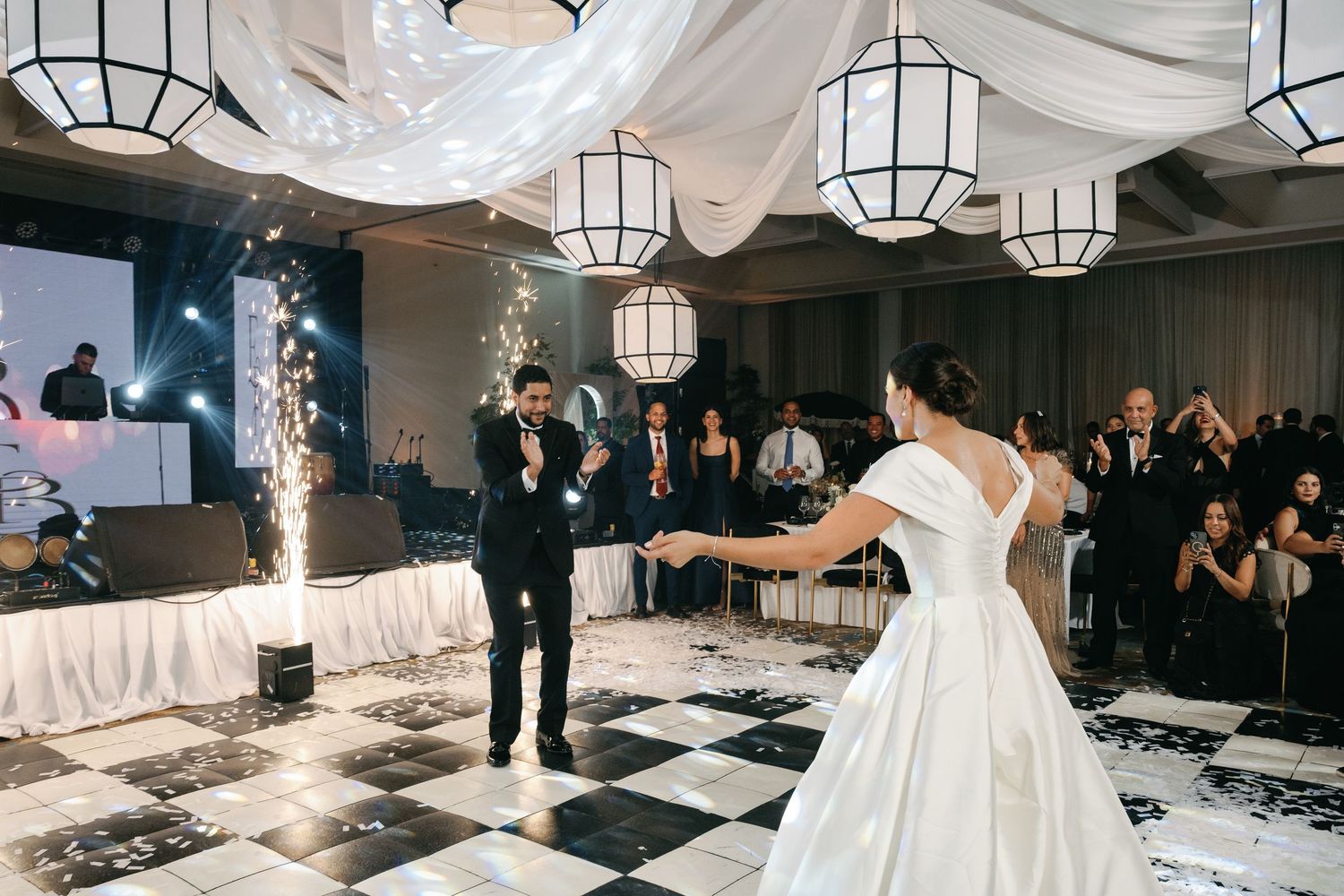 Joyful dancing at a wedding reception on a black and white checkered dance floor with white paper lanterns overhead.