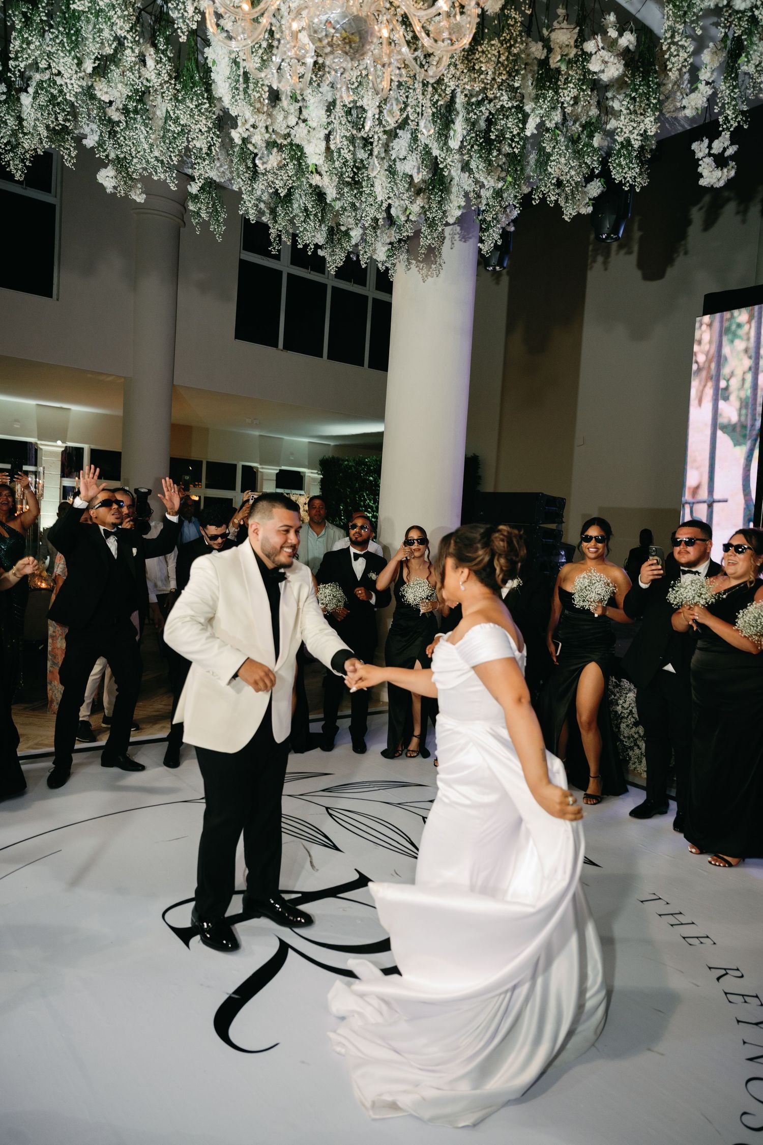 A couple shares their first dance at a wedding reception under hanging floral decorations while guests watch.