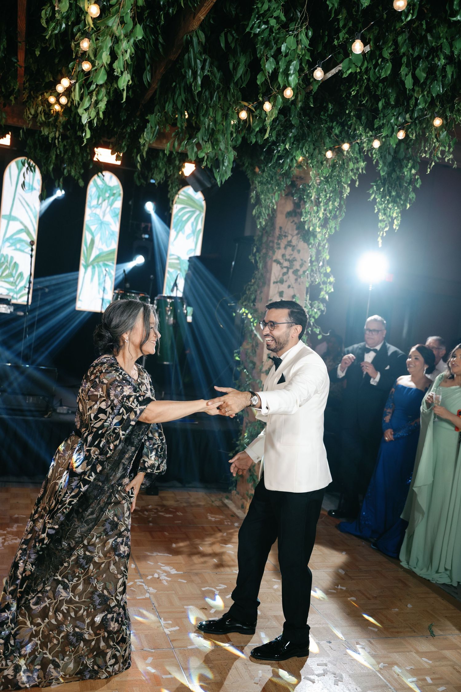 Wedding guests dancing together under decorative lighting and greenery at an indoor reception venue.