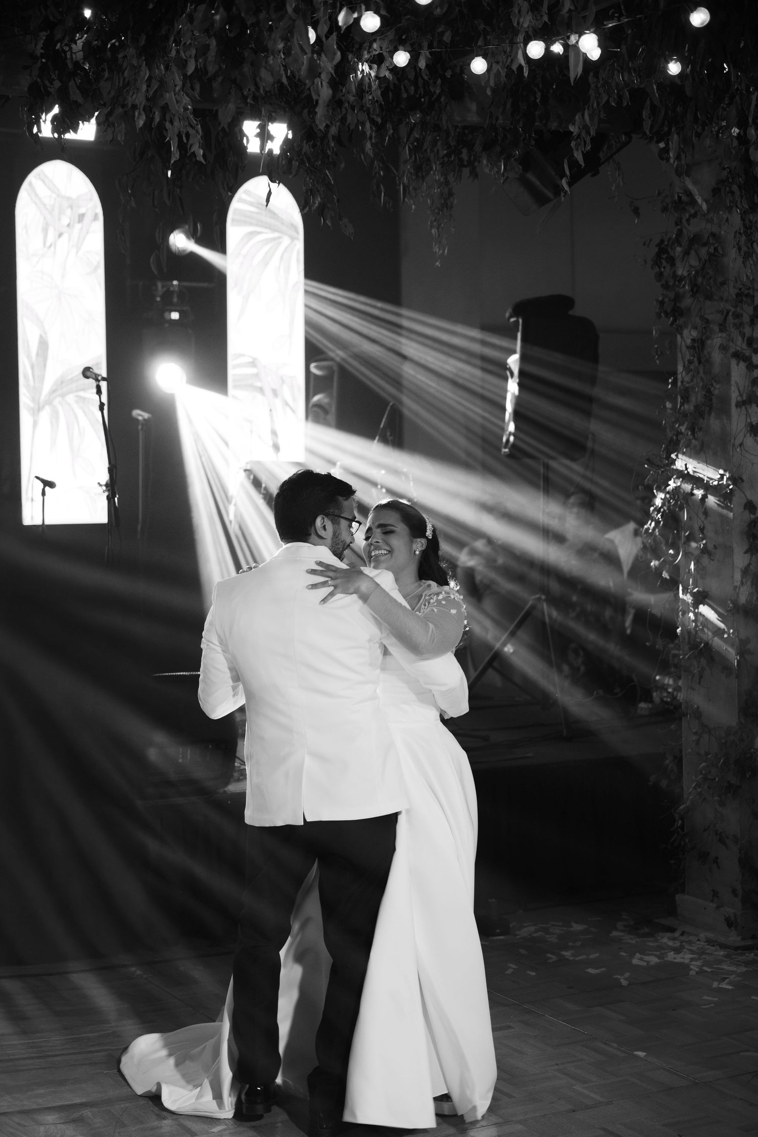 A couple in white attire dances gracefully in dramatic lighting with beams of light streaming through arched windows.