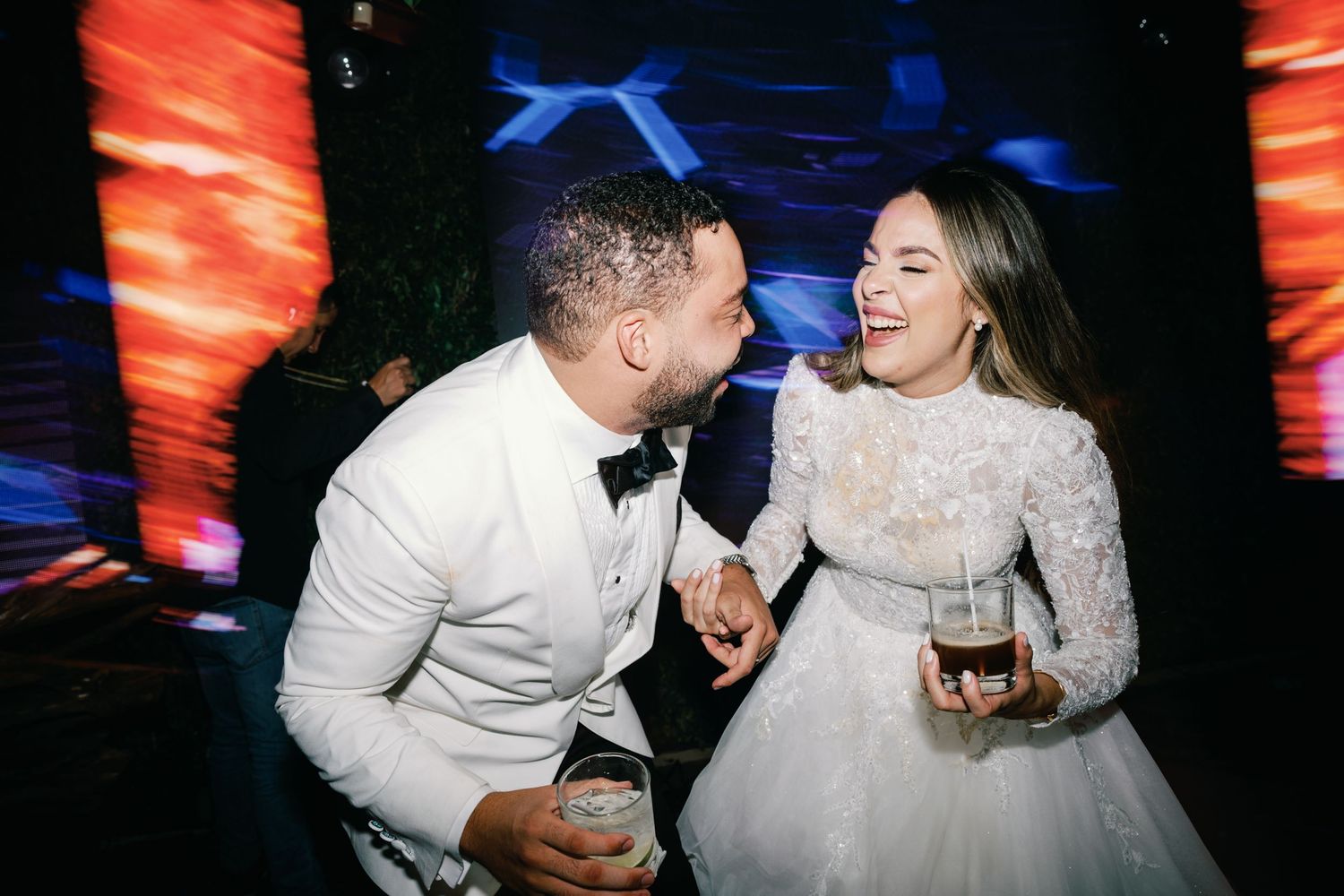 A couple in white wedding attire laugh and dance together at a reception with red and blue lighting effects.