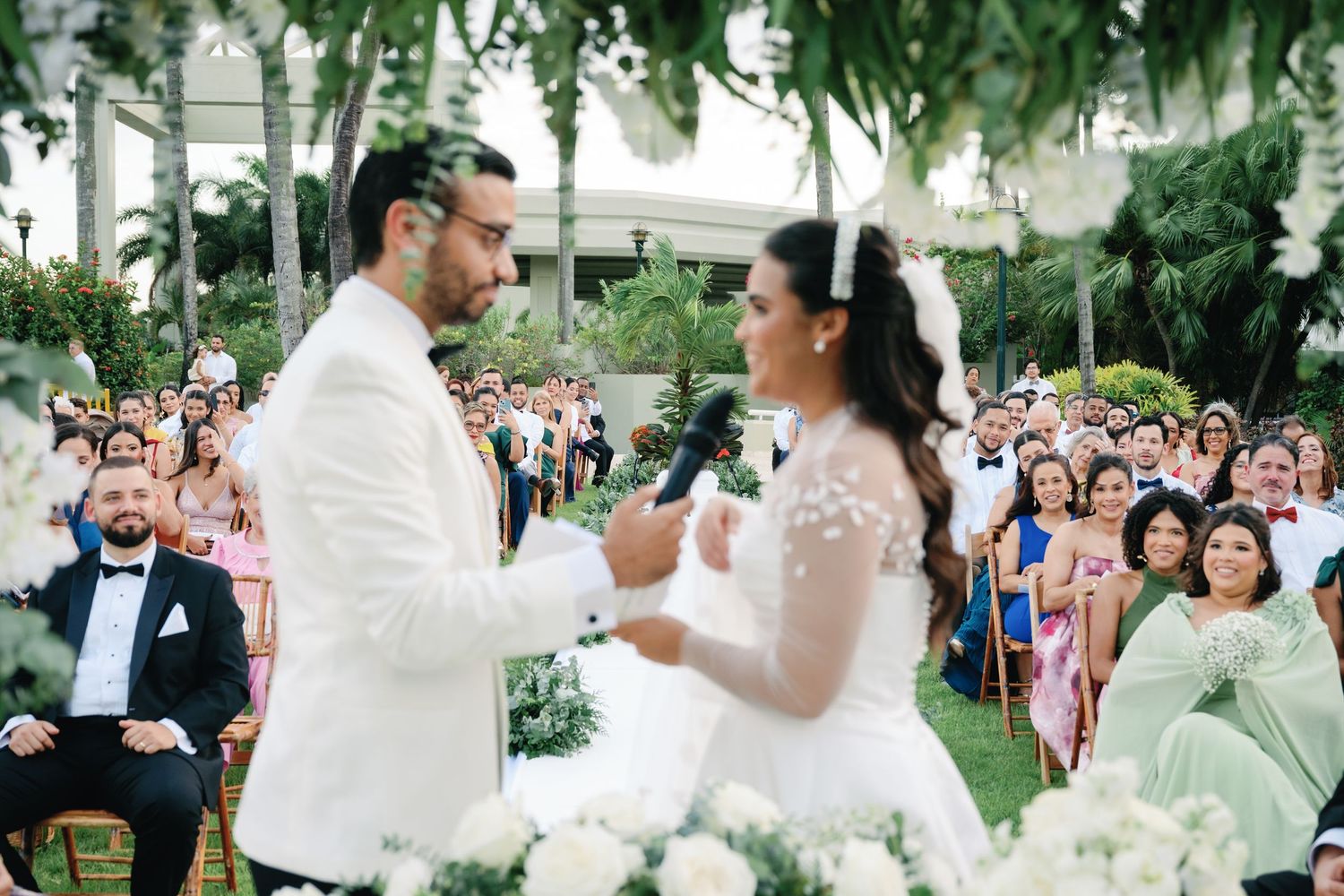 Outdoor wedding celebration with guests gathered under palm trees and string lights at dusk.