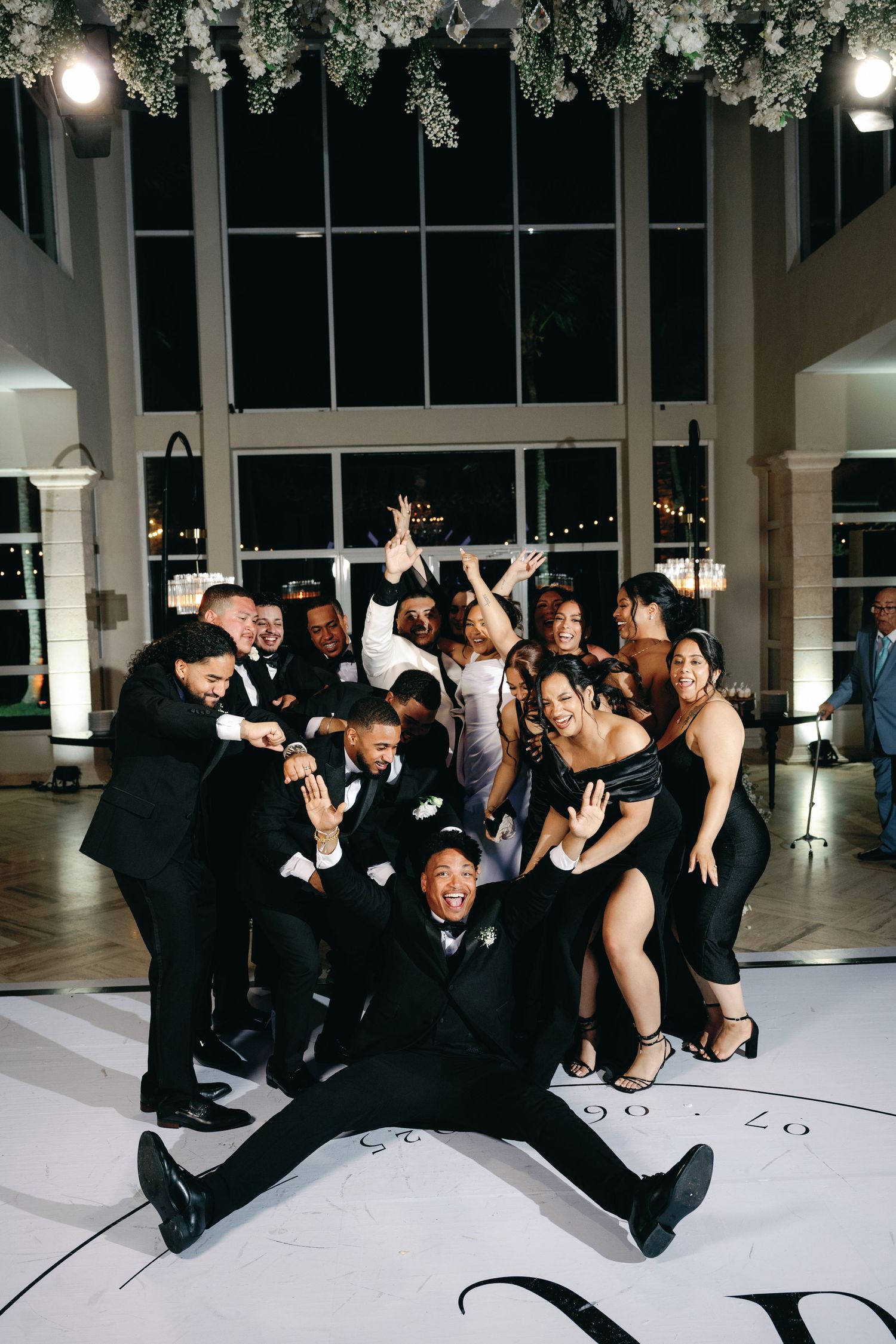 Wedding party in formal black attire poses energetically on a dance floor during an evening reception.