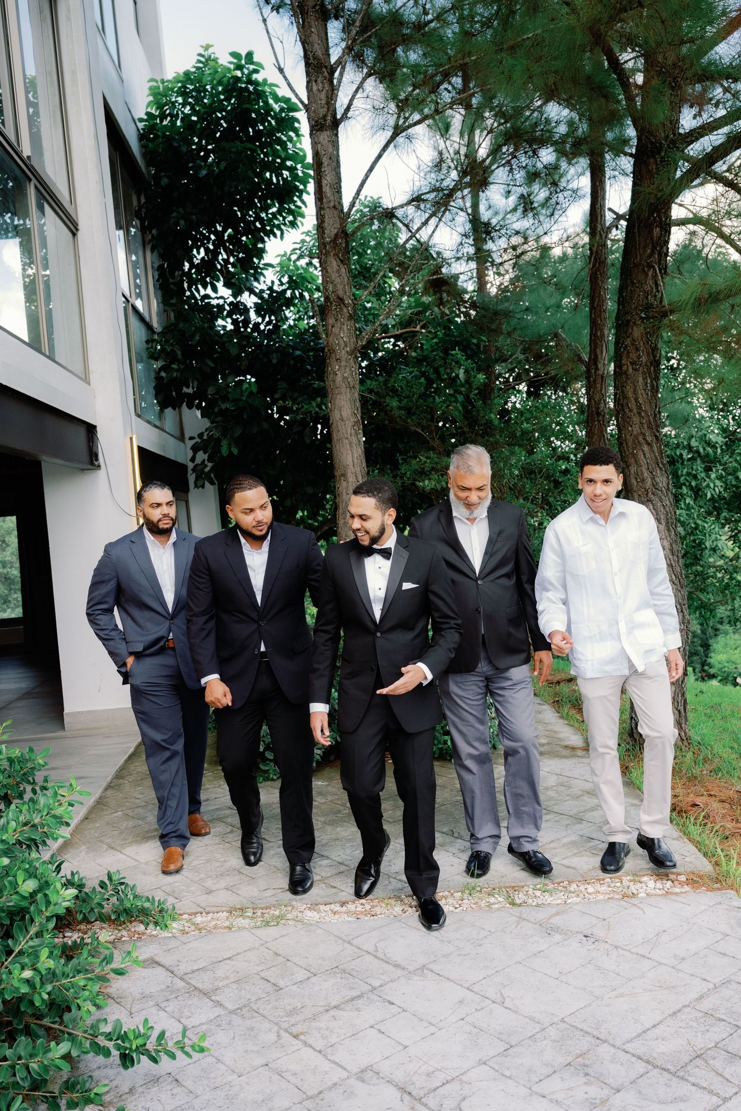 Groomsmen in formal suits walking together on a stone path surrounded by greenery.