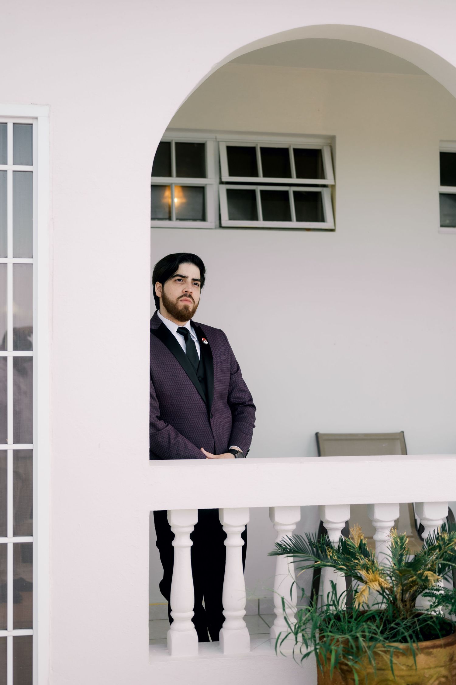 Professional portrait of a person in a dark suit standing on a white balcony with arched windows and potted plants.