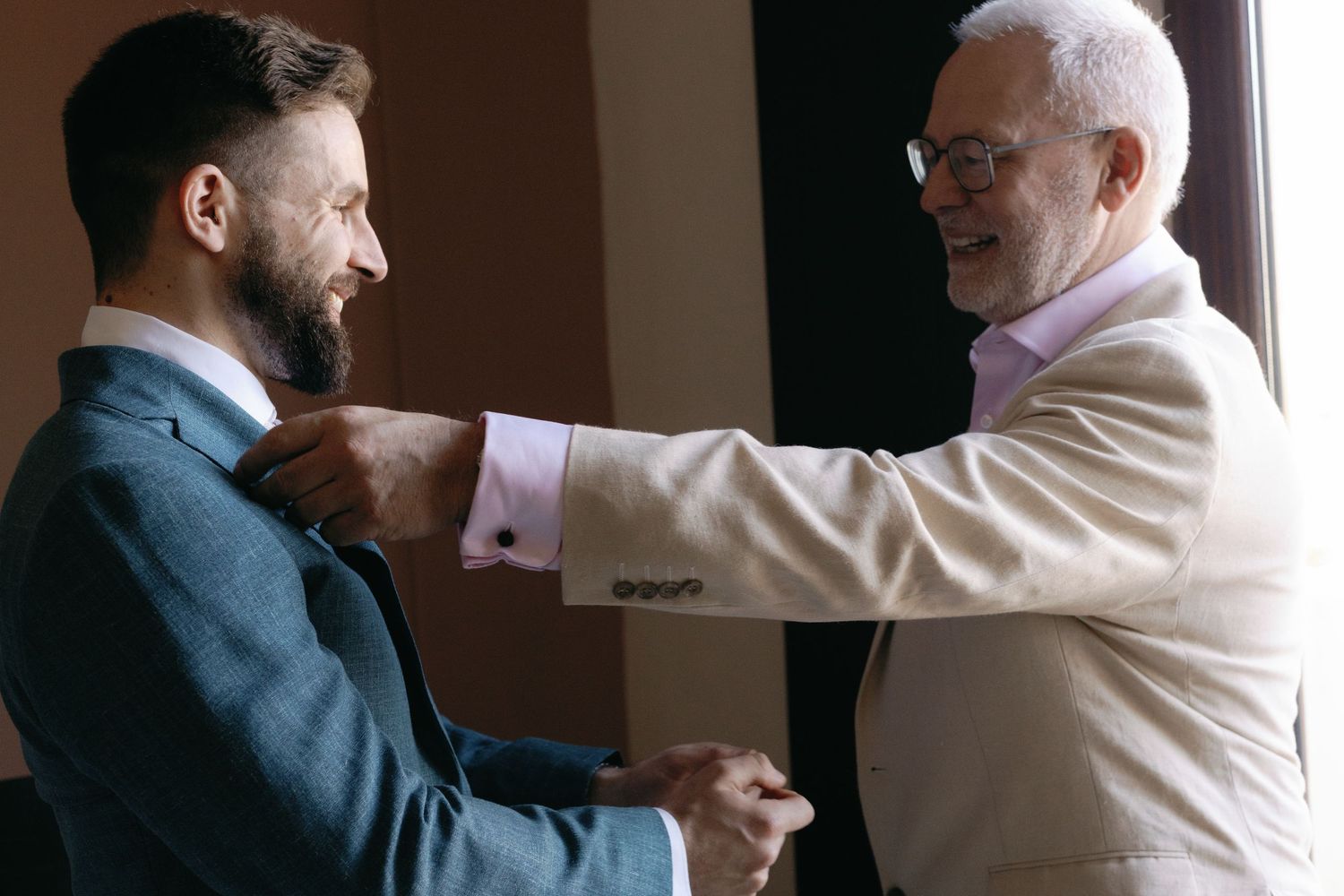 An older man in white helps adjust the tie of a younger man in a navy suit.