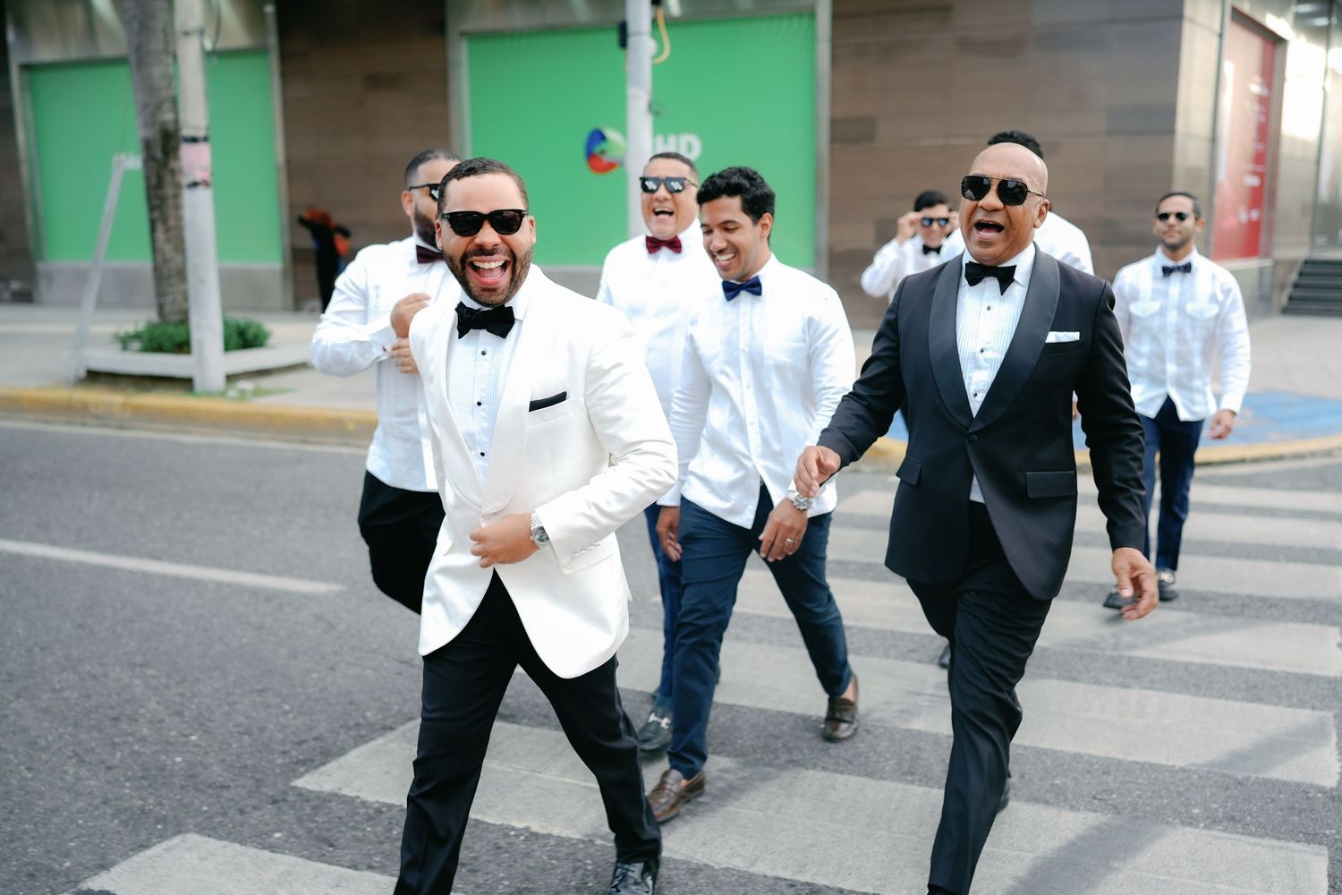 A group of people wearing formal white and black tuxedos walk together on a street with a green wall in the background.