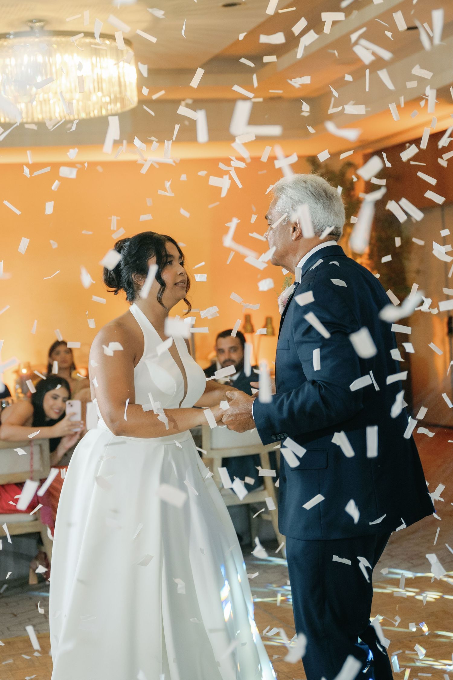Wedding celebration with white confetti raining down as guests dance in a beautifully lit reception hall.