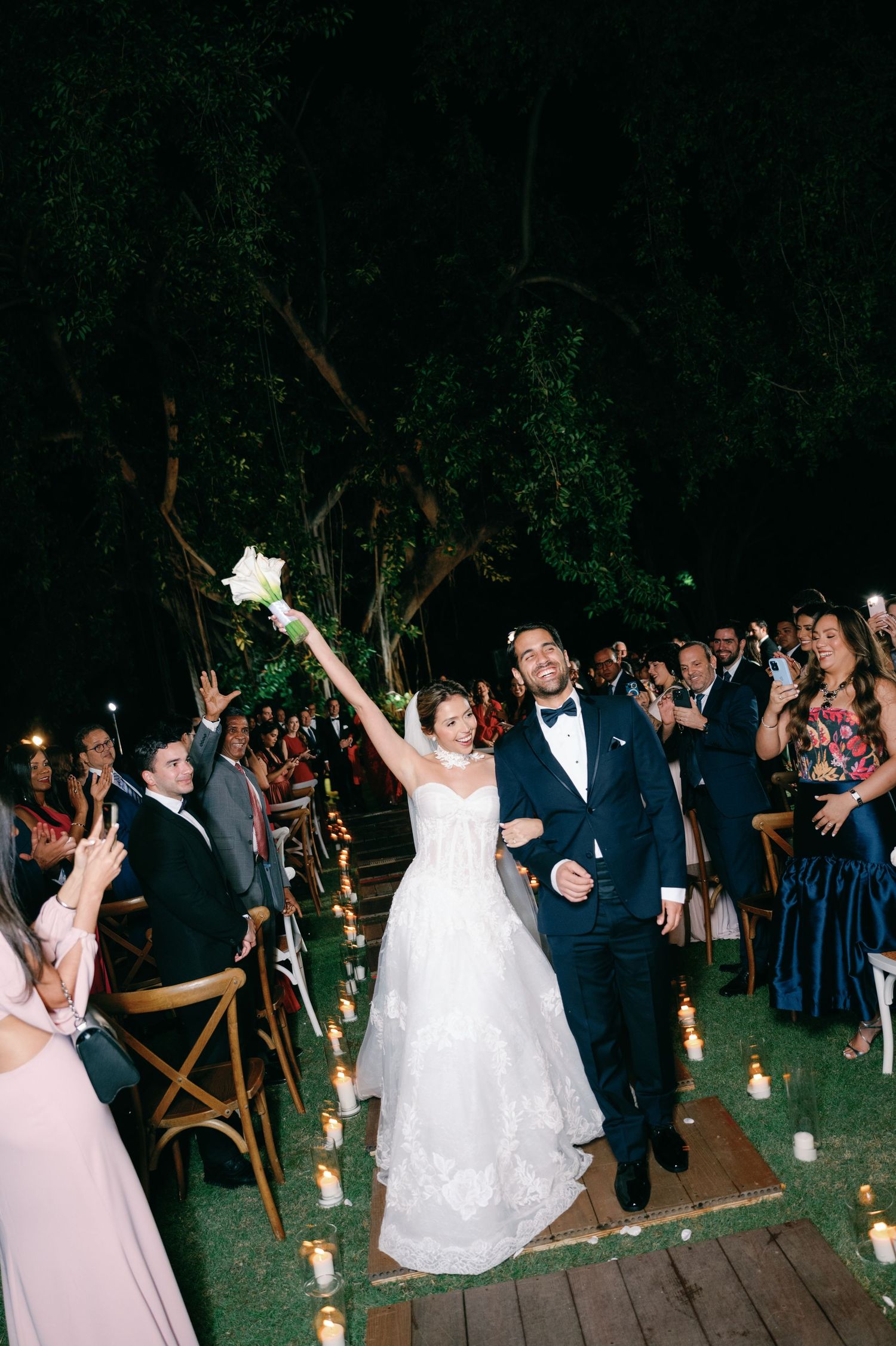 Wedding guests dance and celebrate on a wooden deck surrounded by trees during an outdoor evening reception.