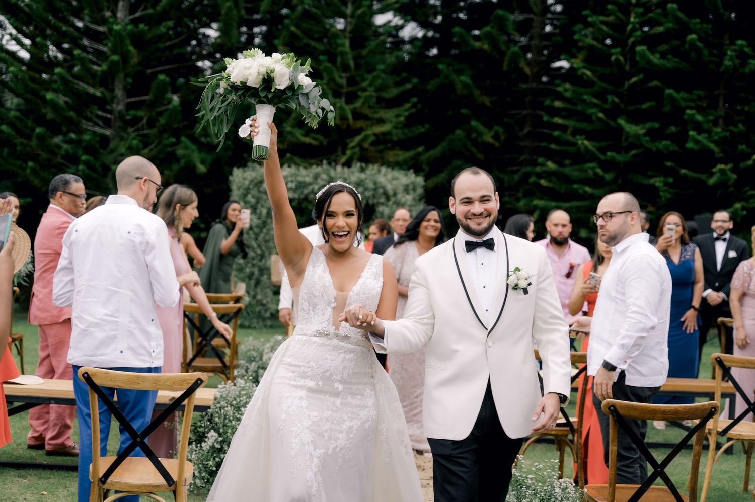 Wedding guests dance and celebrate at an outdoor reception surrounded by evergreen trees in a forest setting.