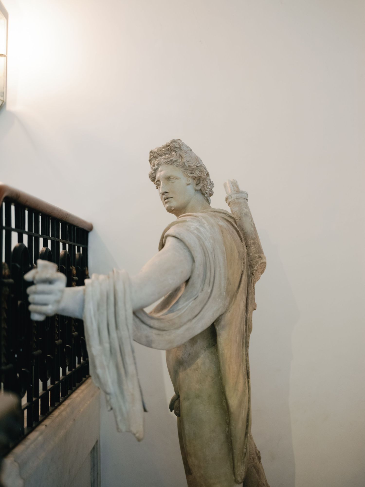 Classical marble statue holding draped fabric stands along an ornate dark staircase railing against a white wall.
