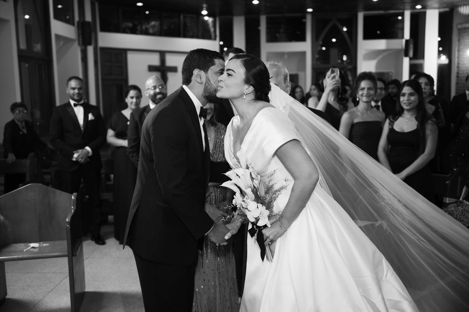 A black and white wedding photograph captures a romantic kiss between newlyweds as guests look on in the background.