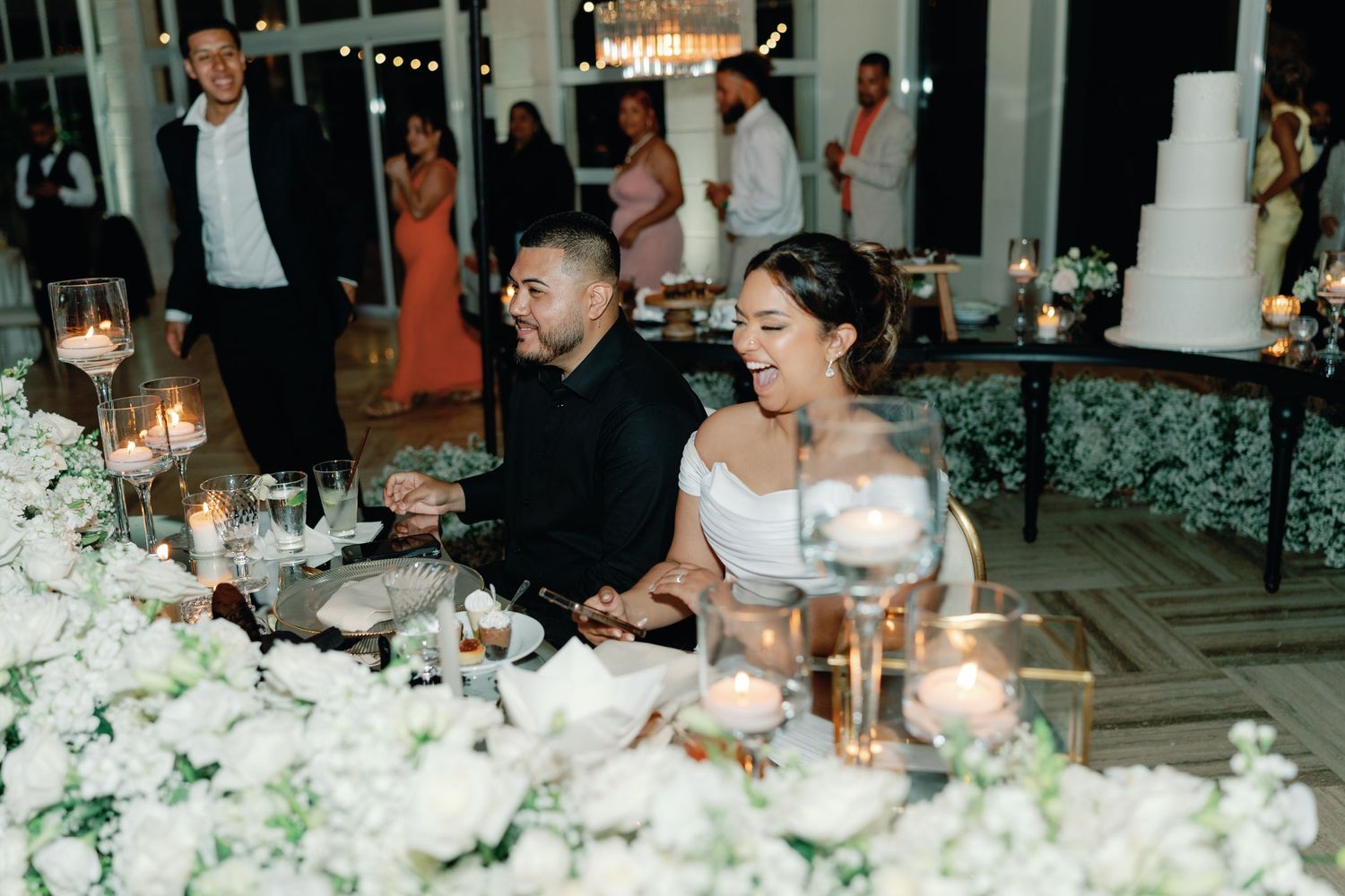 Elegant wedding reception dinner table with white floral centerpieces and glowing candles in a grand ballroom setting.