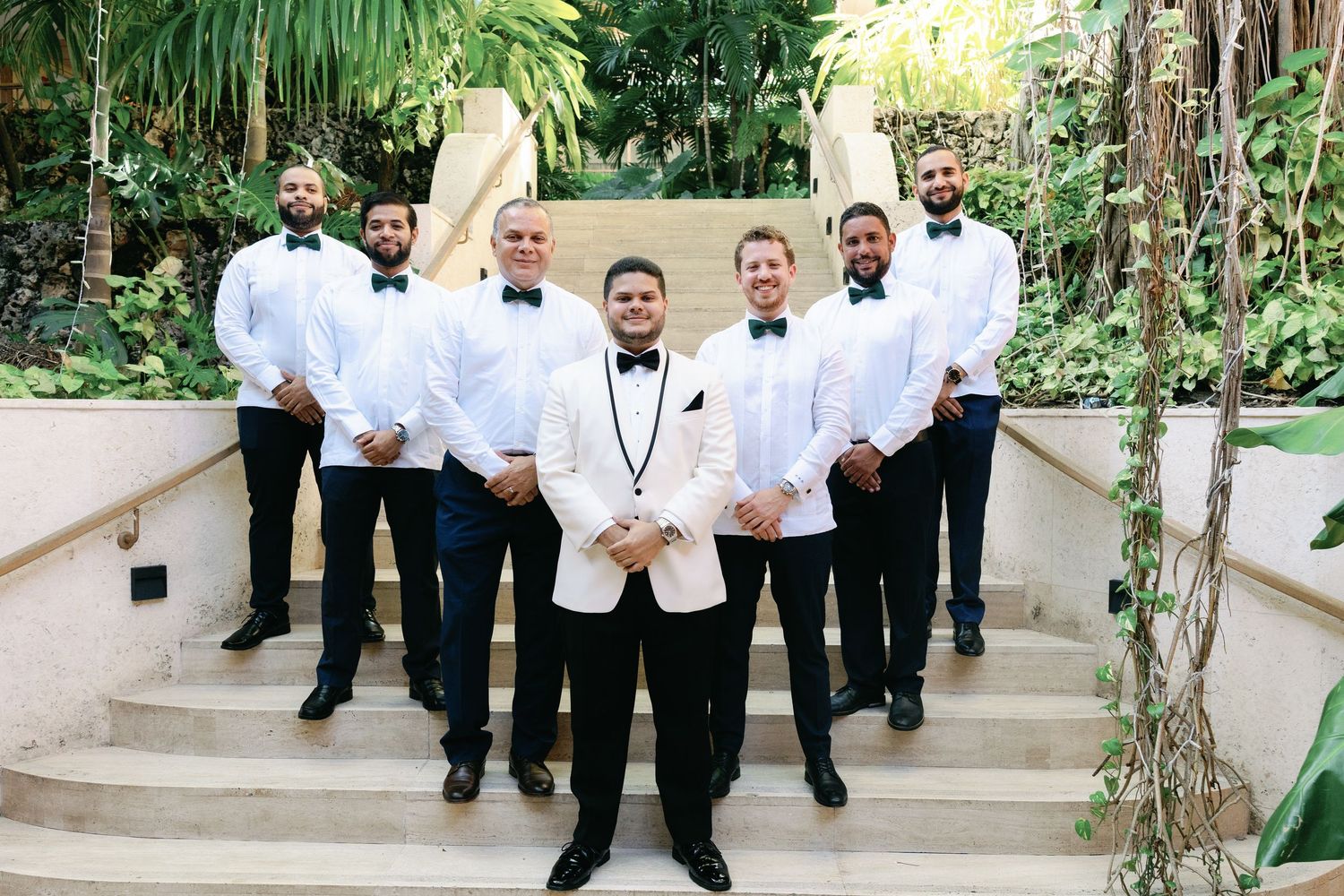 A group of groomsmen in white dinner jackets and black tuxedos pose on outdoor steps surrounded by tropical plants.
