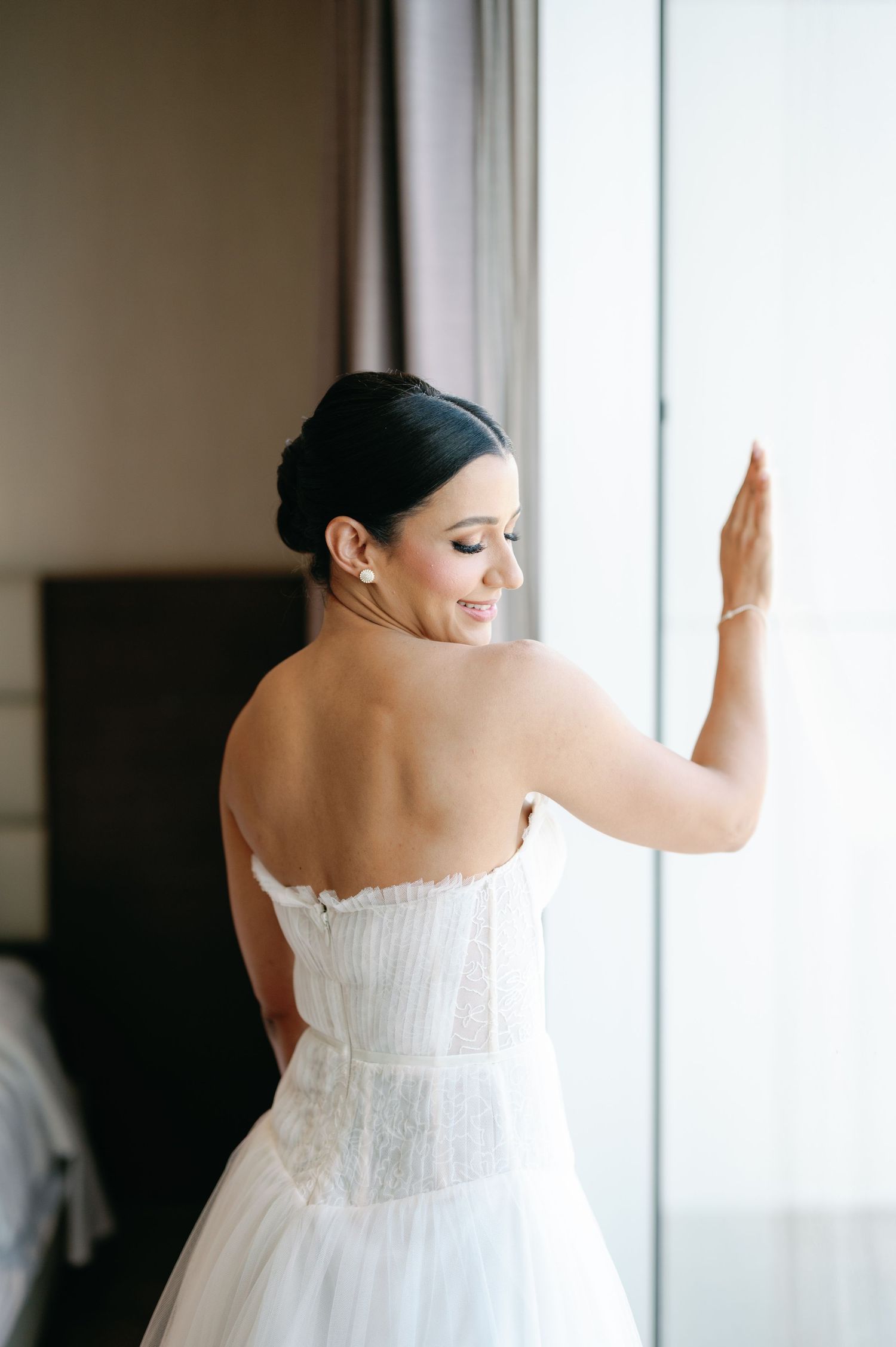 Bride in a white strapless wedding dress leans against a bright window in an elegant indoor setting.