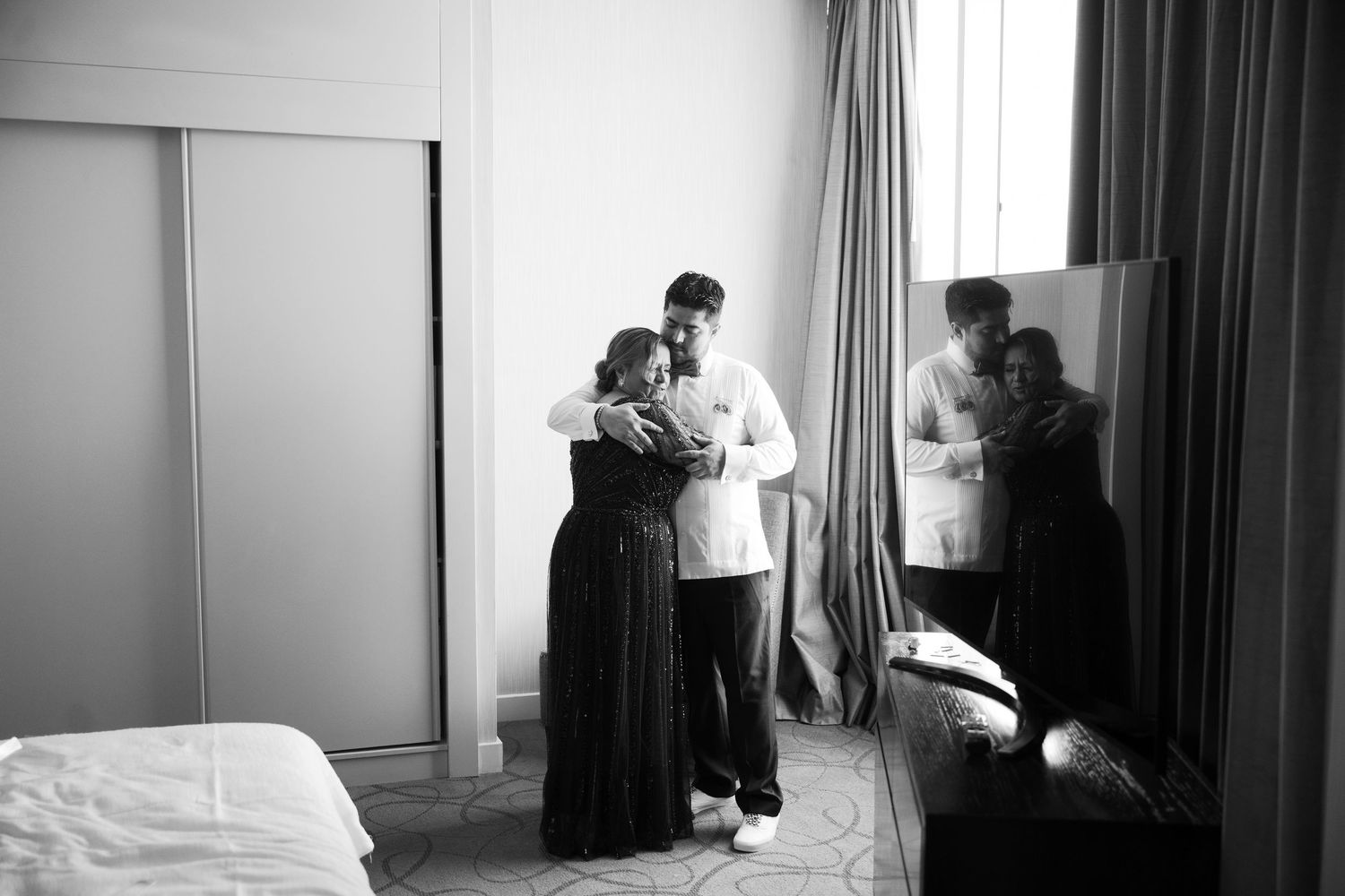 A black and white photograph of people gathered in a bedroom, with curtains and sliding doors visible in the background.