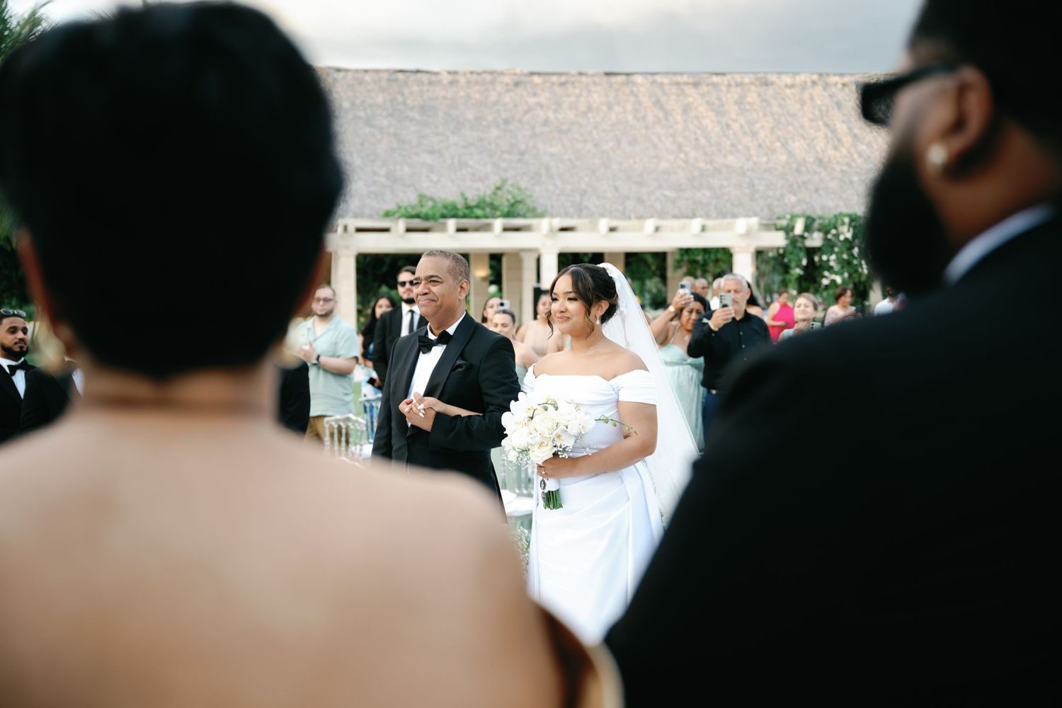 A wedding ceremony takes place under a white tent canopy with guests gathered around.