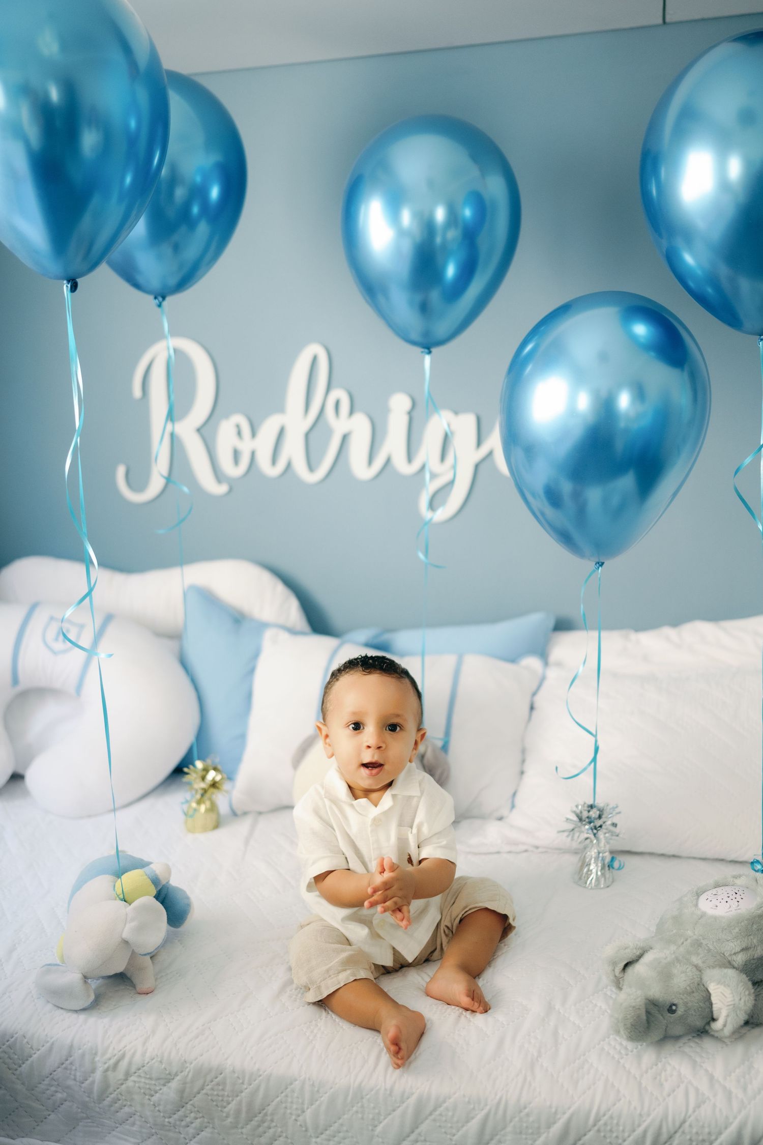 Baby boy named Rodrig sits on white bed surrounded by blue metallic balloons and stuffed animals for birthday celebration.