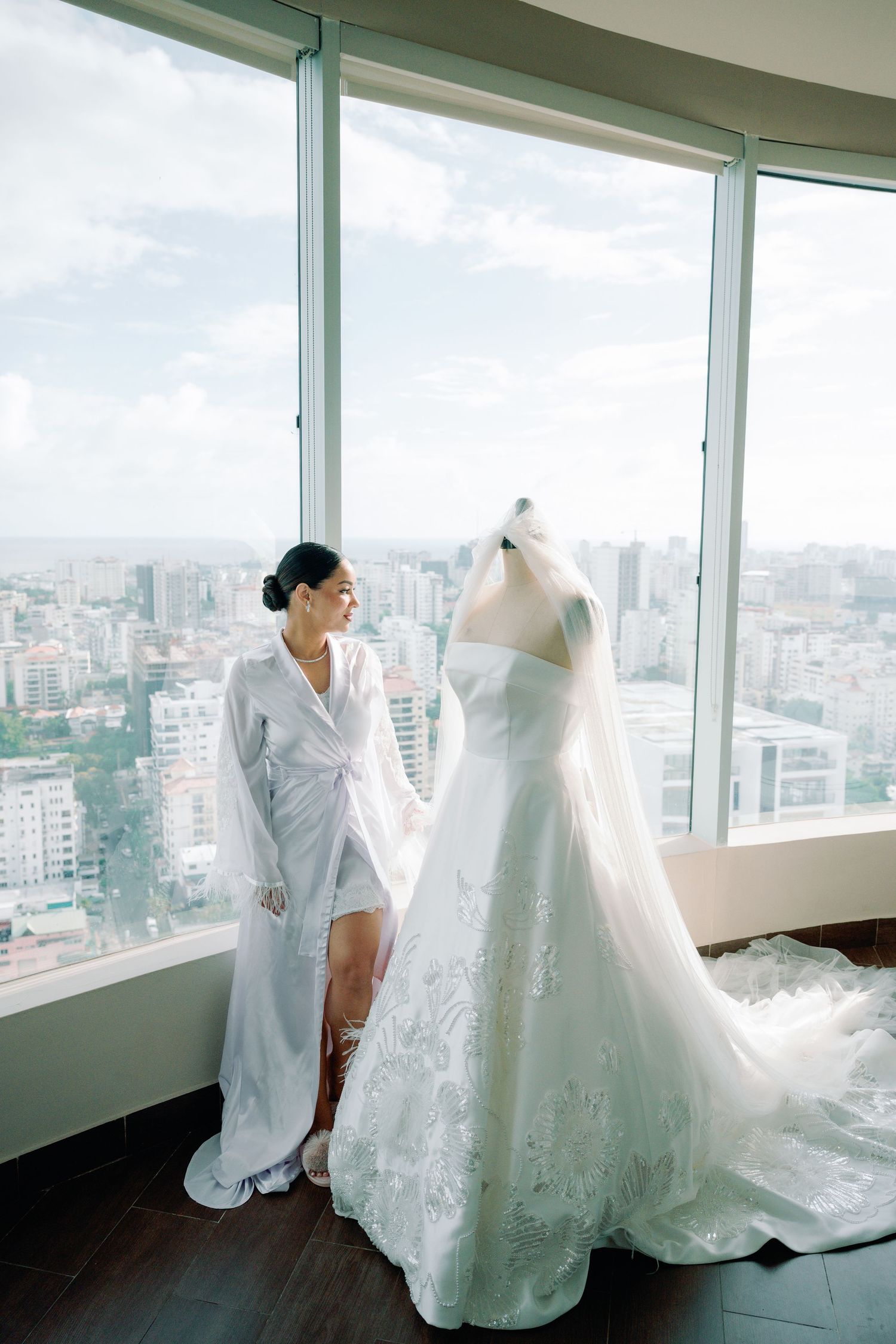 Wedding dress display in a high-rise room with floor-to-ceiling windows overlooking a cityscape.