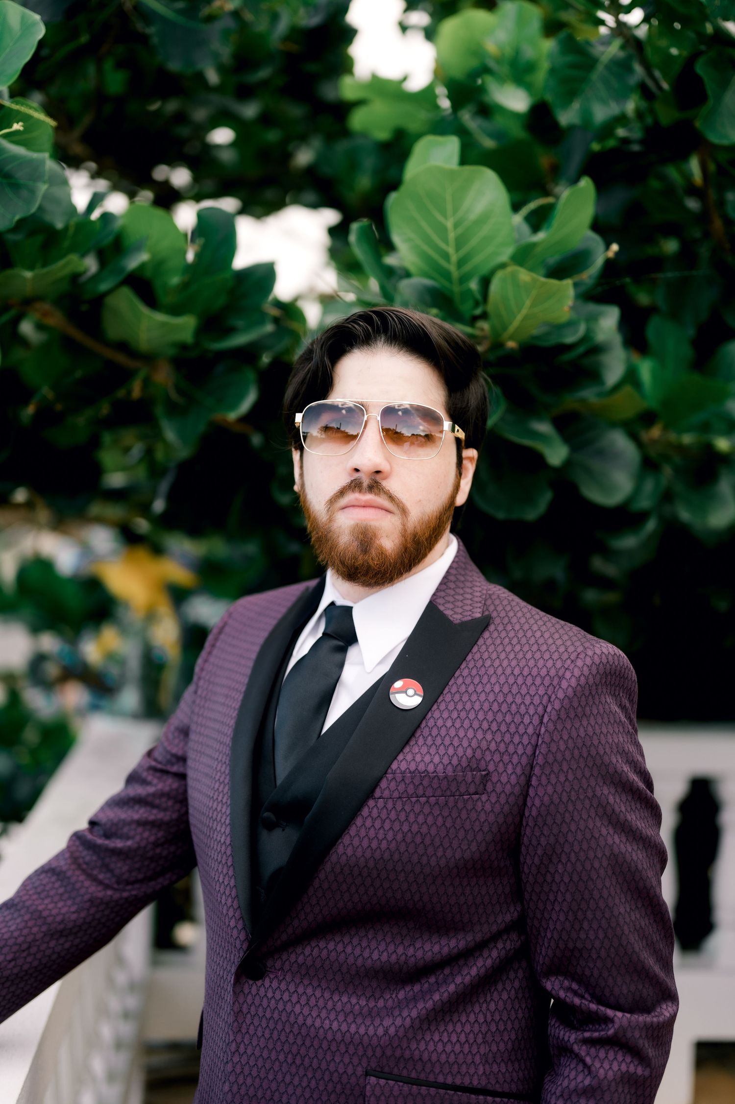 Person wearing purple patterned suit jacket and sunglasses stands confidently against leafy outdoor backdrop.