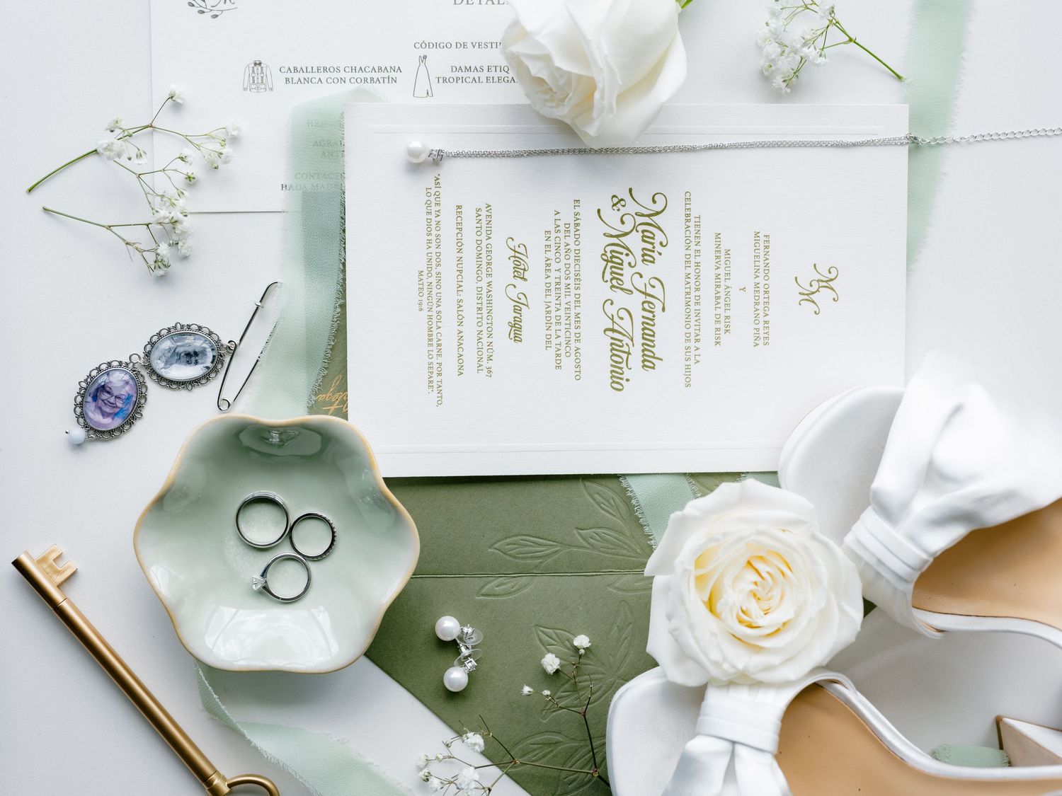 Elegant wedding flatlay with white roses, rings, and stationery on soft sage green and white background.