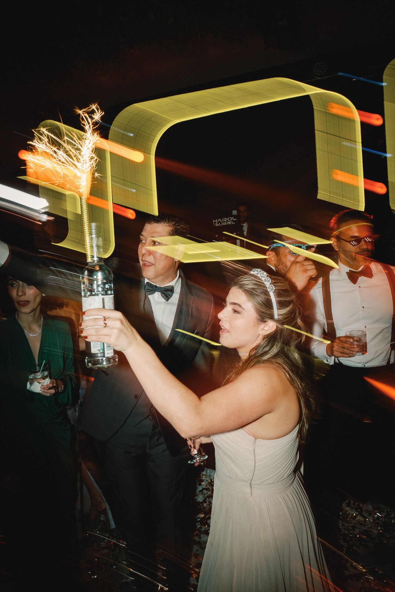 Wedding guests dance and celebrate with sparklers under yellow lighting at an indoor reception venue.