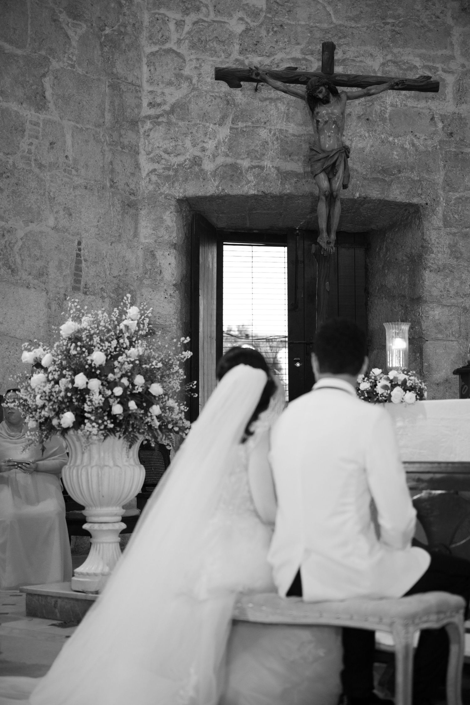 A black and white wedding scene shows a couple kneeling at an altar in a stone church with a crucifix on the wall.