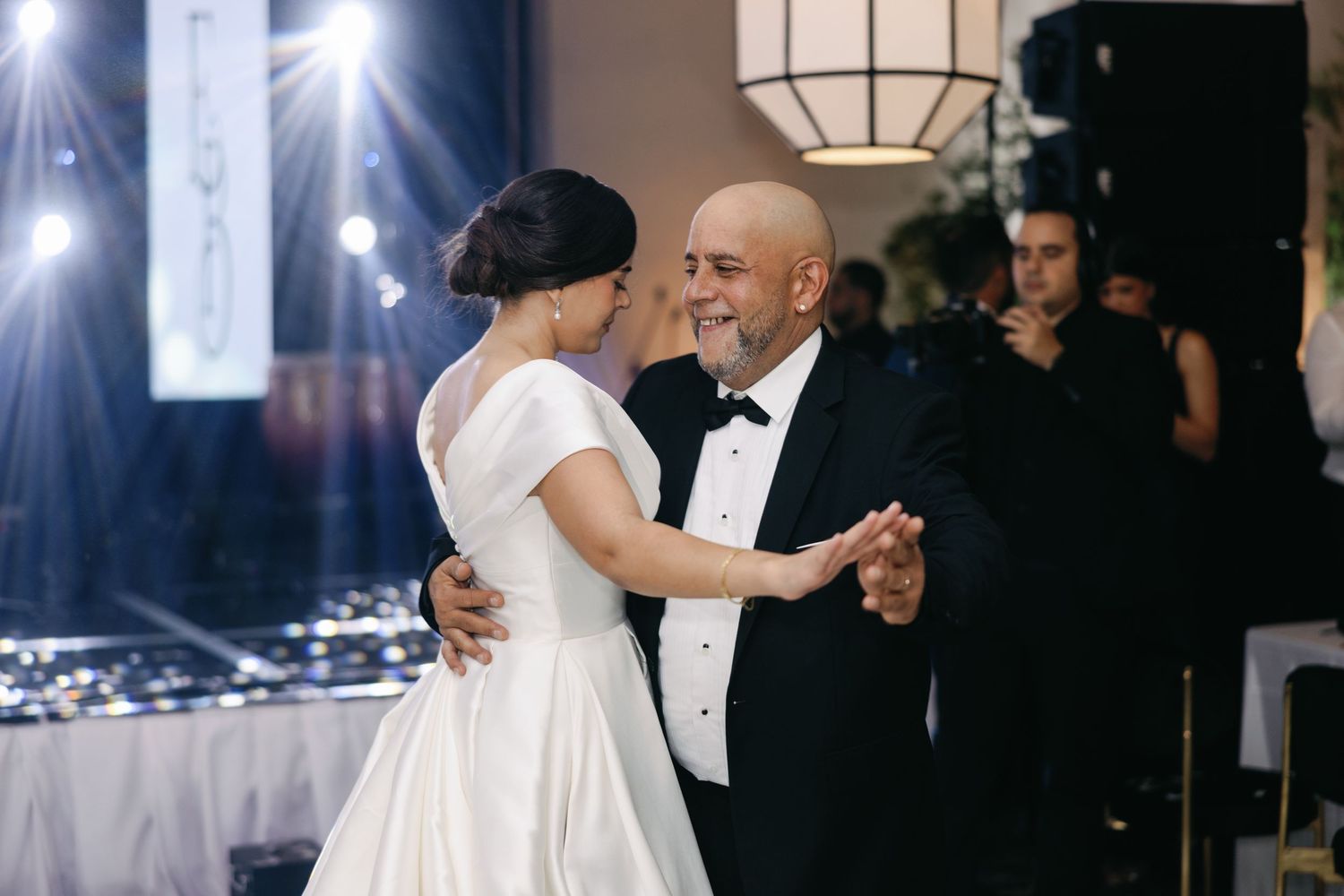 A couple in formal wedding attire dance together at an elegant reception with dramatic lighting and guests in the background.
