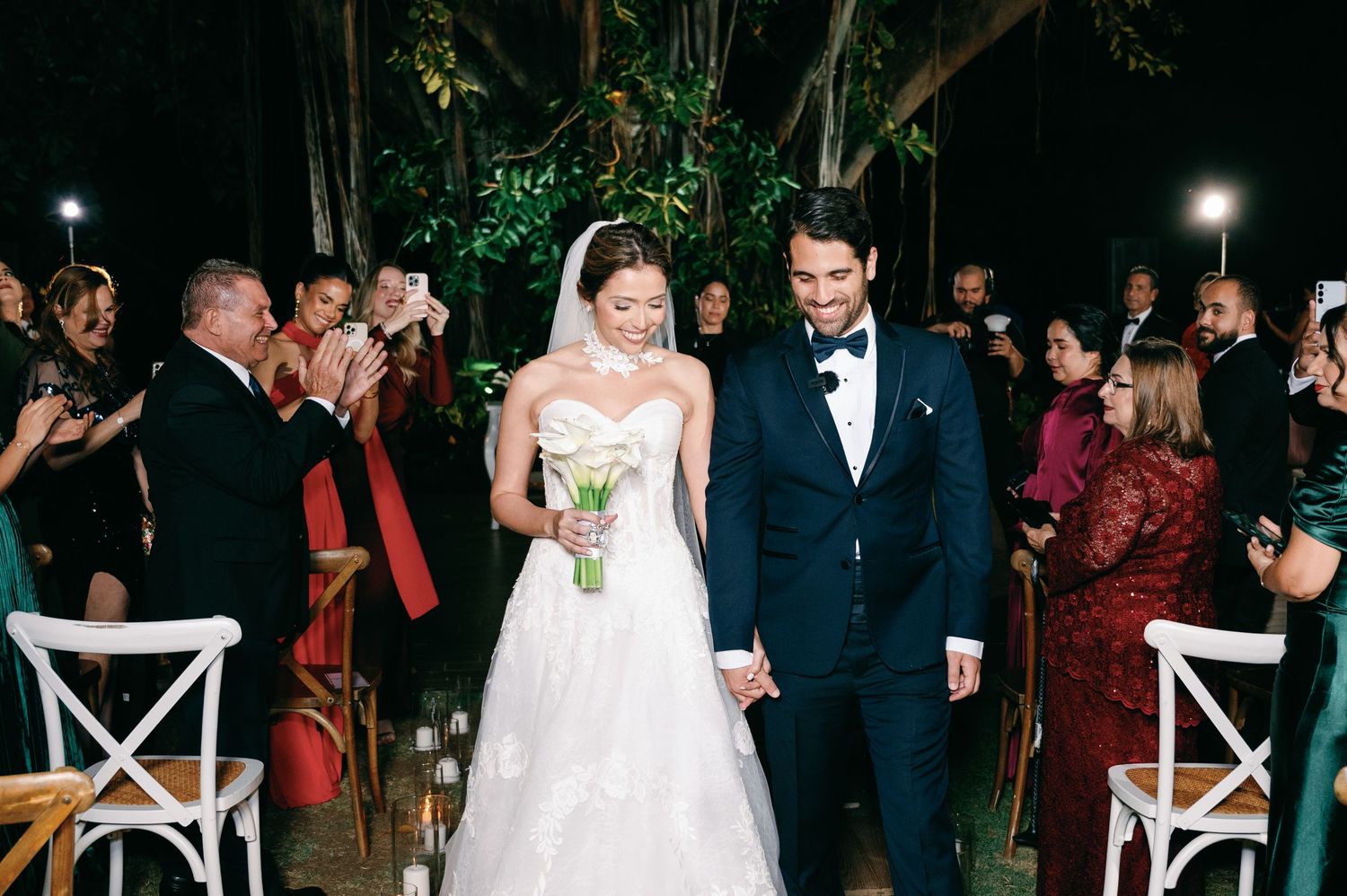 Couple walking down the aisle at an outdoor evening wedding ceremony with white chairs and string lights.