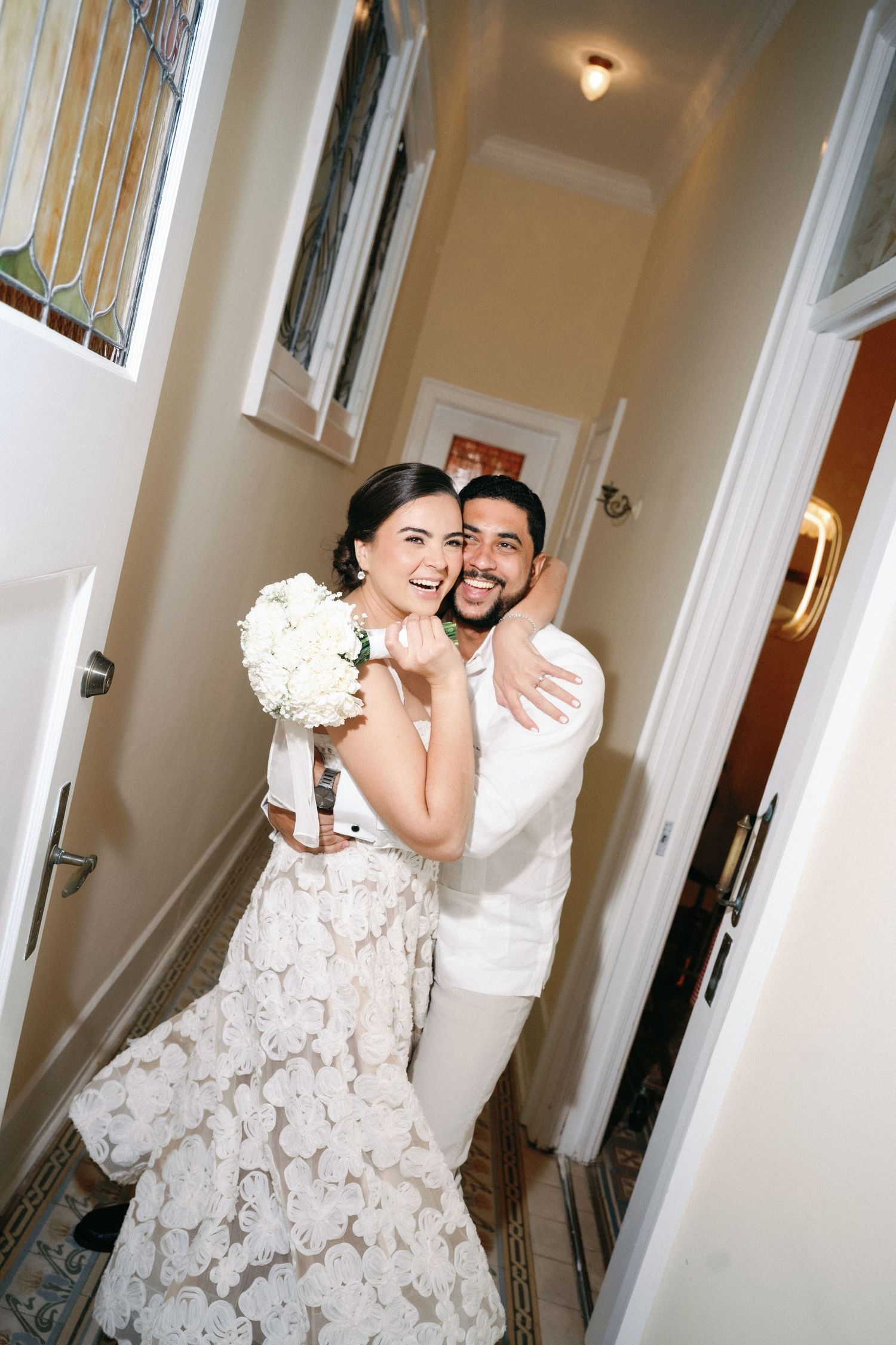 A couple in wedding attire shares a joyful moment in a bright hallway with white walls and wooden doors.