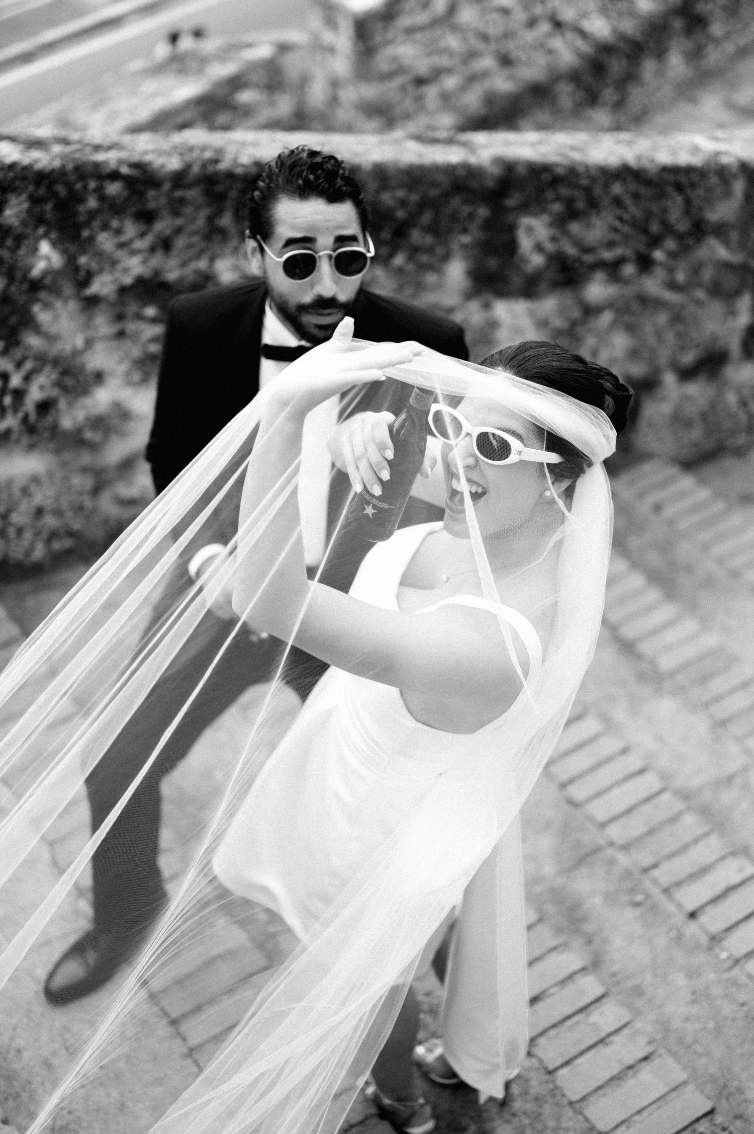 A couple in sunglasses playfully posing with flowing white fabric on a brick pathway.